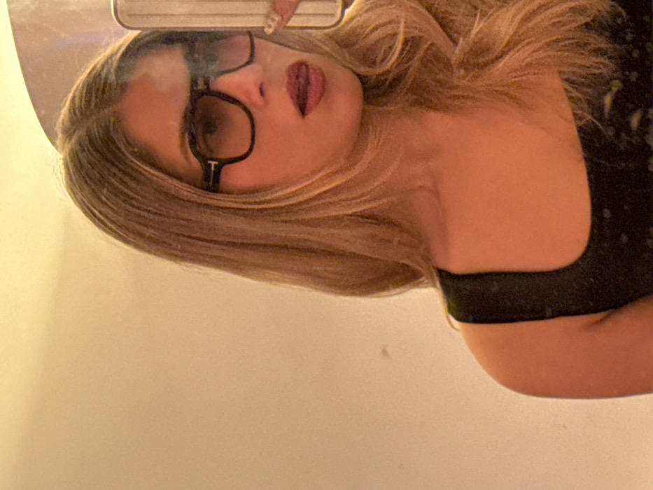 A woman with long blonde hair, wearing glasses, poses in front of a mirror. She wears a dark top with thin straps. The reflection captures her face, showing her lips slightly parted and eyes looking directly at the camera. The mirror's edge and a partial view of a phone can be seen. The background is a plain, light-colored wall.