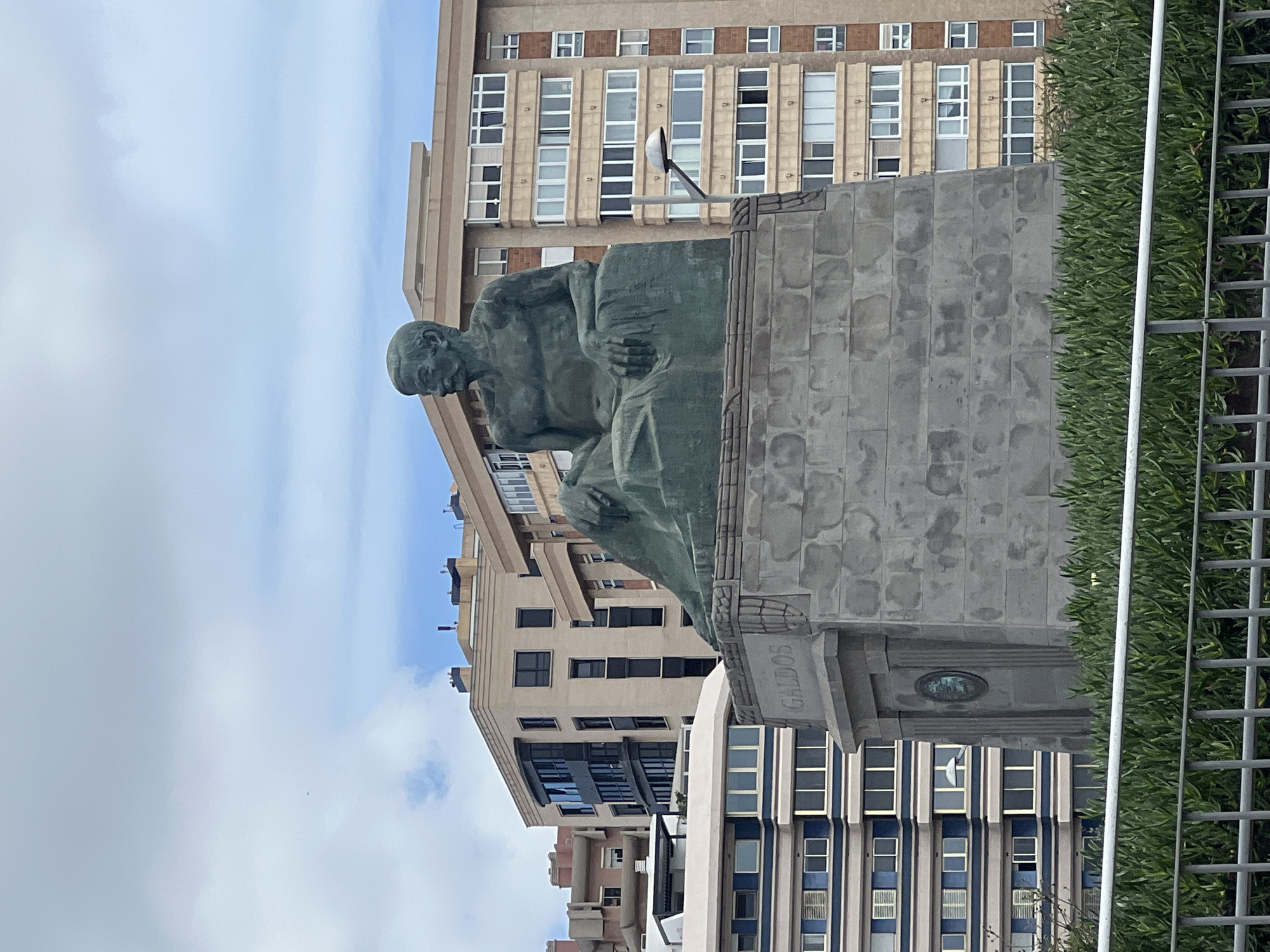 The image shows the statue of Benito Pérez Galdós, a prominent Spanish novelist, playwright, and politician, located in Las Palmas de Gran Canaria, Spain. The statue depicts Galdós seated in a contemplative pose. It is mounted on a stone pedestal, which has the name "GALDOS" inscribed on it. The buildings of Las Palmas form a background, with an apartment building visible behind the statue. Metal fencing is present on the right. The sky is partly cloudy.