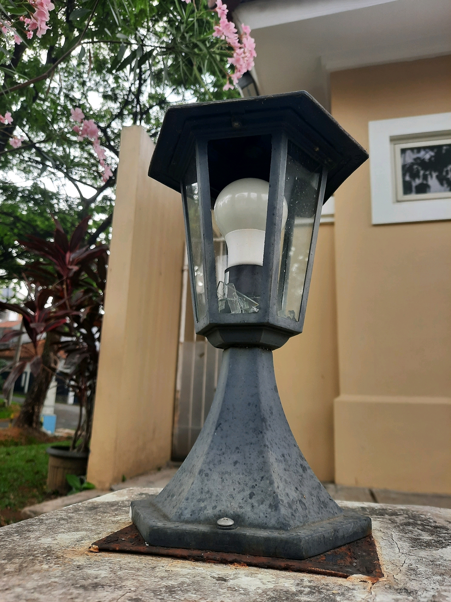 In Babakan Madang, Indonesia, an outdoor lamp post with a broken glass panel stands on a textured concrete surface. The lamp has a dark gray, hexagonal base and a metal frame holding a white light bulb. In the background, a beige building with a white-framed window is visible. Lush green foliage and delicate pink flowers from a nearby tree frame the top left of the image. The overall scene suggests an outdoor setting with a touch of nature.