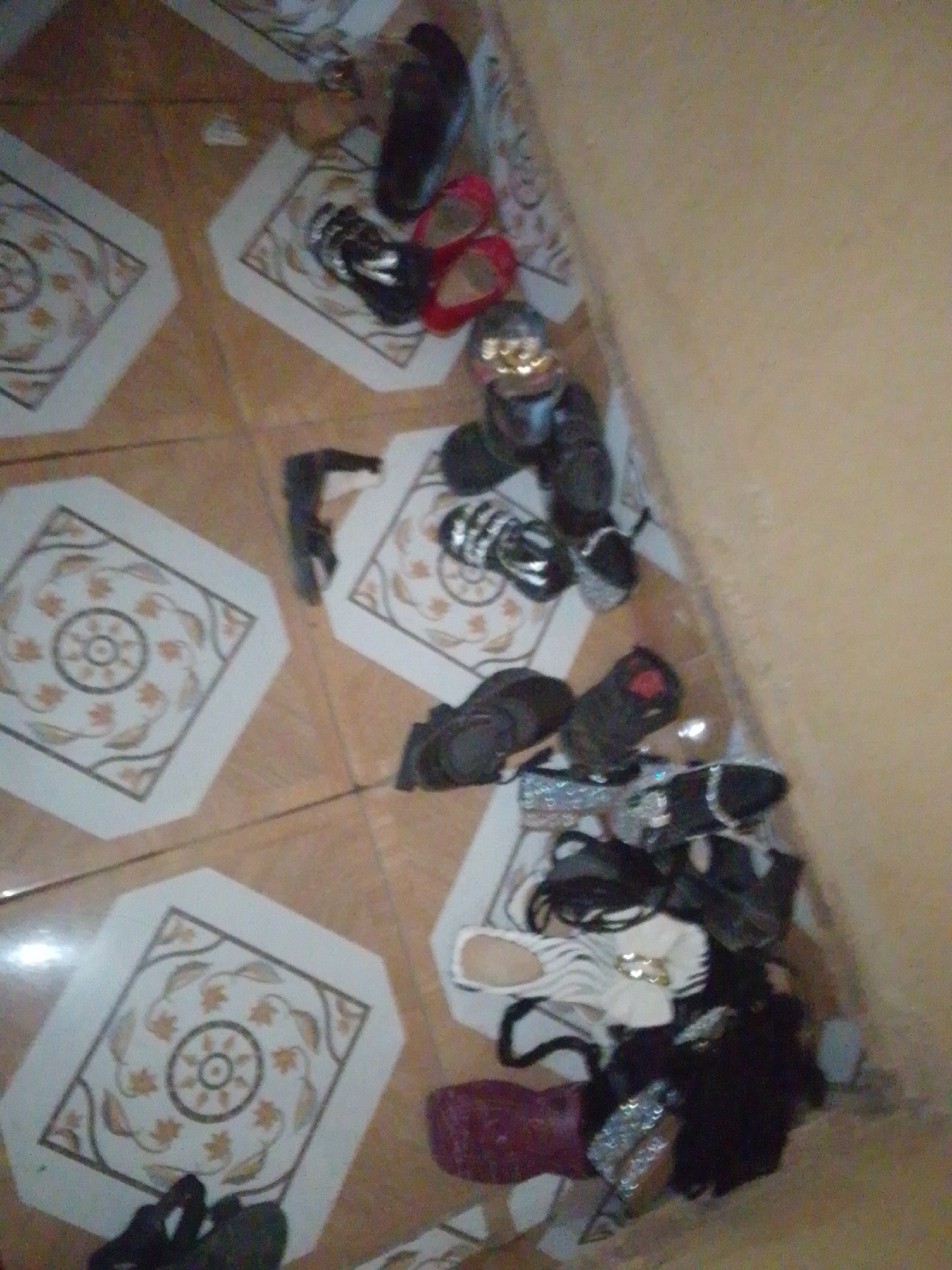 The image shows a collection of shoes scattered on a tiled floor. The tiles are light brown with octagonal inserts featuring a decorative pattern of leaves and a circular motif. Several pairs of shoes are visible, including black high heels with sparkly embellishments, red sandals, black flip-flops, and some black and white sandals. One dark-colored boot is also present. The shoes are arranged in piles, some in the foreground and others further back. A light-colored wall is visible on the right side of the image, and the lighting suggests an indoor setting, possibly a home or a shop. There is no indication of people or any activity taking place, other than the shoes being placed on the floor. The overall scene appears to be a disorganized arrangement of footwear. The location is identified as Jalingo, Nigeria.