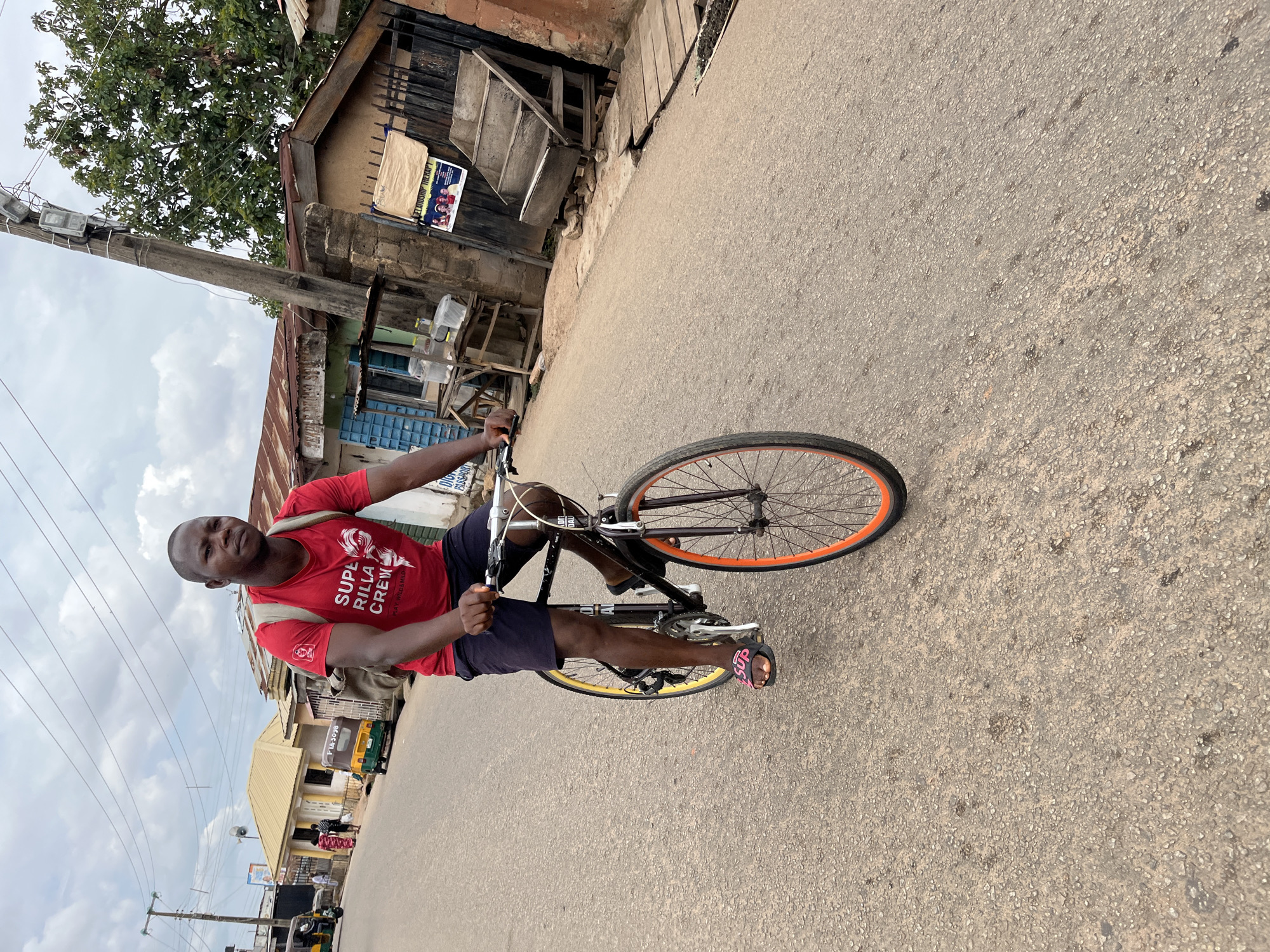 A male individual is observed cycling a black bicycle with a yellow rear wheel and an orange front wheel, along a paved road in the city of Jos, Nigeria. The man, appearing to be in his late 20s to early 30s, wears a red t-shirt with the text "SUPA RILLA CREW" and a graphic of a gorilla's head, black shorts, and black "Sup" branded slide sandals with pink lettering. A light-colored backpack is visible on his back. He maintains a forward gaze towards the viewer while pedaling.
The road surface is asphalt, exhibiting minor wear and scattered debris. On the left side of the road, a yellow-roofed building is visible amidst other structures with corrugated metal roofs. Two auto-rickshaws (locally known as Keke Napep), one green and one yellow, are parked or moving slowly. Several utility poles with multiple overhead electrical wires traverse the scene against a partly cloudy sky. Further down the road, figures of pedestrians are discernible.
On the right side of the road, closer to the foreground, a concrete utility pole with multiple light fixtures and numerous electrical wires stands prominently. Beyond the pole, residential or commercial structures with corrugated metal roofs are present. A wooden table with plastic containers is positioned near a building with blue-barred windows. A brown cardboard sign displaying "RELATIONSHIP THERAPY" with images of people is affixed to a metal gate. A wooden stepped structure, possibly for display or seating, is also visible. Green foliage from a tree extends over the buildings on the right. The overall environment suggests a typical urban street scene in Nigeria.