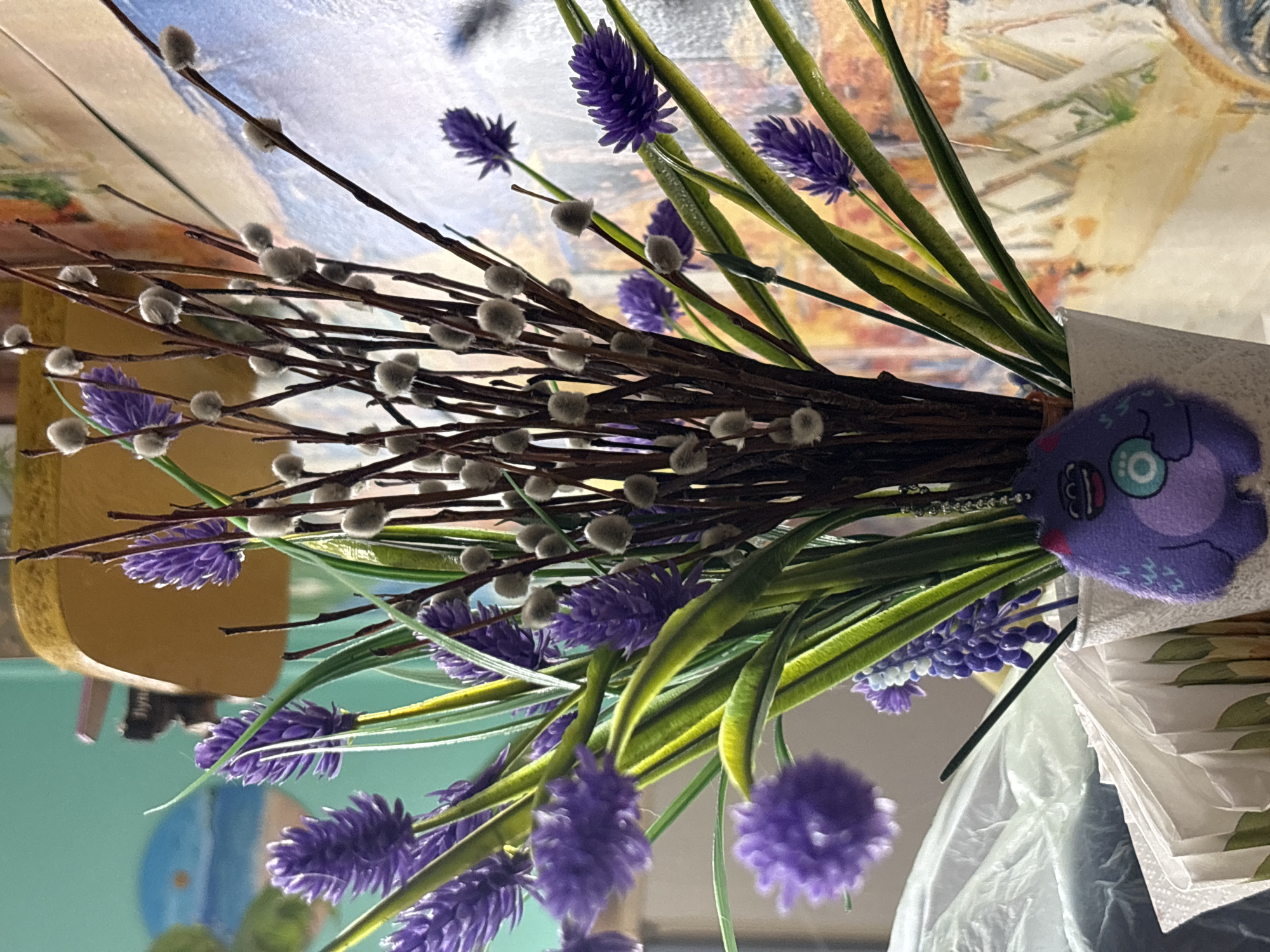 The image shows a bouquet of flowers consisting of pussy willow branches and purple hyacinth-like flowers. A small purple plush monster toy is tucked in among the flowers near the base. The flowers are in a white paper or plastic wrapping. The background is blurry but appears to be a painting or a wall. There is no information in the image to suggest that this is happening in the city of Марковский, Russia.