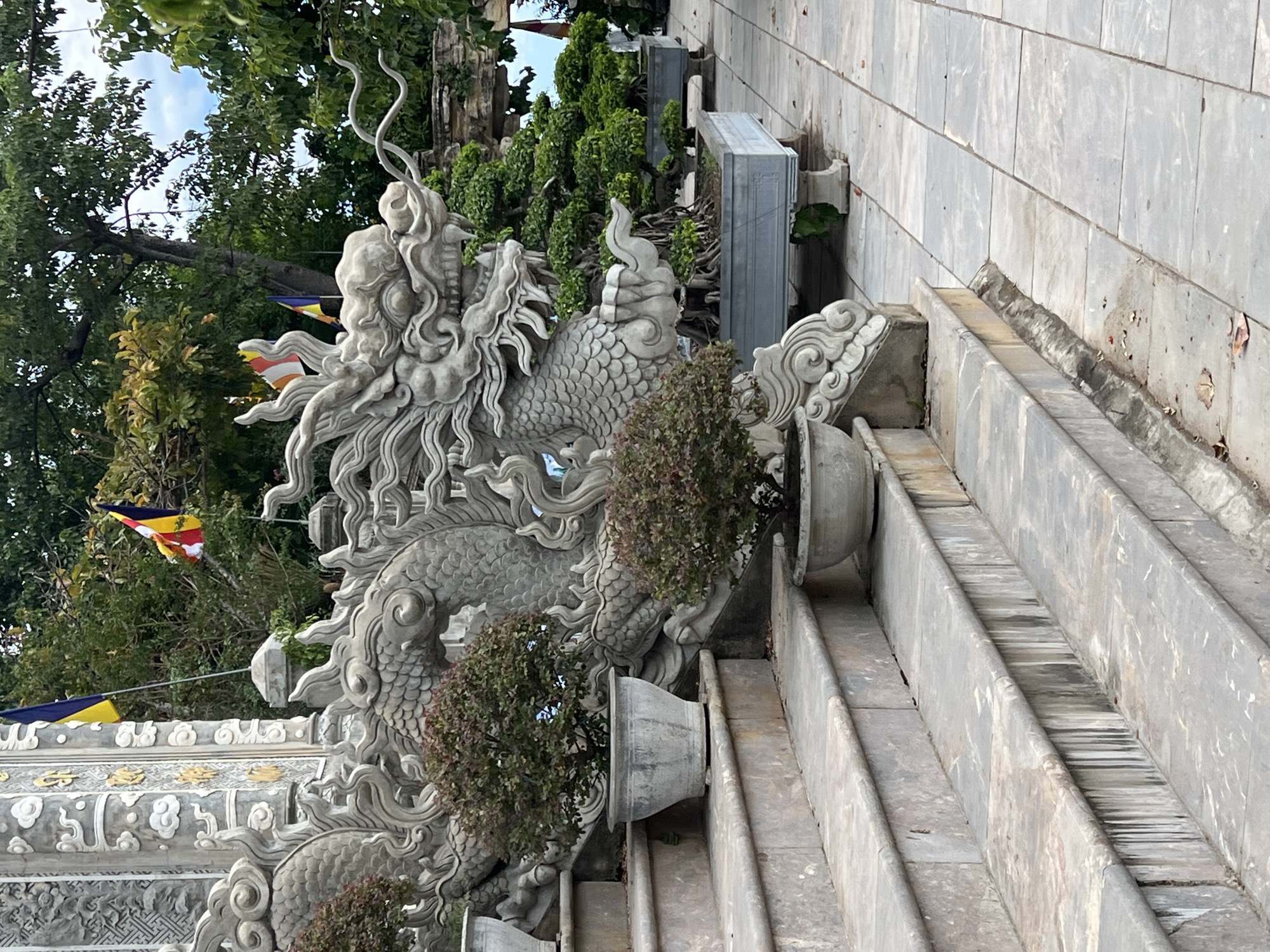 The image shows a stone dragon sculpture at the entrance of a building, likely a temple or pagoda, in Da Nang, Vietnam. The sculpture is intricately carved with scales, a mane, and whiskers, and it appears to be guarding a set of stone steps. Potted plants, including bonsai trees, are placed along the stairs and in front of the sculpture. In the background, there are more trees and what appear to be Buddhist flags. The overall scene suggests a tranquil and spiritual setting, possibly within a temple complex like Linh Ung Pagoda on Son Tra Peninsula, which is known for its large Buddha statue and dragon-themed architecture. The paving stones and the weathered stone of the dragon sculpture indicate an outdoor location, exposed to the elements. No people are visible in the image.
