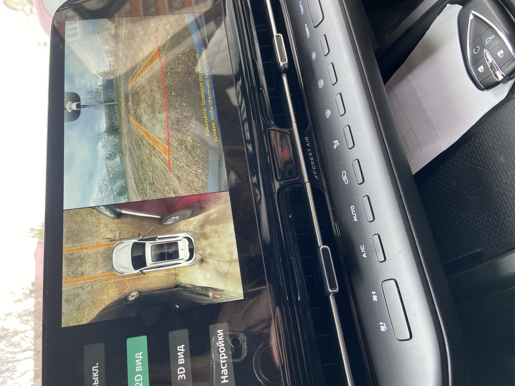 The media file captures the interior of a vehicle, specifically focusing on its central display screen and adjacent dashboard controls. The scene is set inside a modern car, likely in Chaikovsky, Russia, given the provided context and visible Cyrillic text.
The primary subject is the car's infotainment screen, which displays a parking assist or 360-degree camera system. The screen is split into two views:
1. The upper portion shows a rear-camera perspective looking backwards over a dirt or gravel path, with dynamic guiding lines (red and orange) overlaid. Distant buildings and vegetation are visible in the background. A warning message in Russian, "Пожалуйста, проверьте окружающие условия, чтобы обеспечить безопасность!" (Please check the surrounding conditions to ensure safety!), is displayed.
2. The lower portion presents a top-down, 360-degree view of the car, depicted as a white SUV, parked or maneuvering between other vehicles (a red car to the upper right and a black car to the lower left). Below this view are interactive buttons labeled "Выкл." (Off), "2D вид" (2D view), "3D вид" (3D view), and "Настройки" (Settings), with "2D вид" highlighted, suggesting the current mode.
To the right of the screen, a section of the dashboard is visible, featuring air conditioning and ventilation controls. These include buttons for "TEMP," "AUTO," "A/C," fan speed adjustments, and defroster functions, along with text like "FOREST AIR." A red hazard light button is also present. In the bottom right corner, a modern car key with various remote functions (lock, unlock, trunk release) rests on a textured surface.
The image appears to be taken during the daytime, with ambient light illuminating the car's interior and the screen's display.
