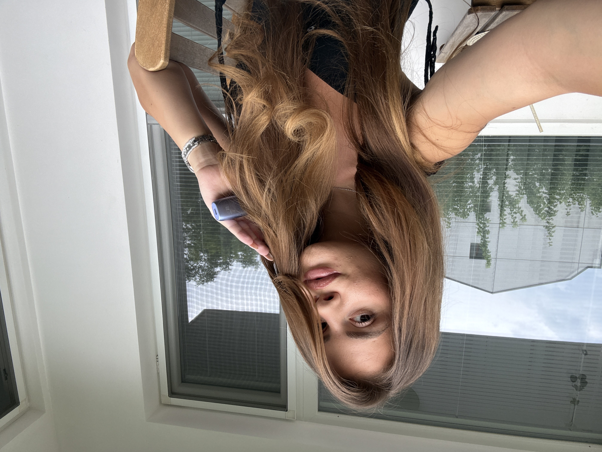 A young woman with long, wavy, brown hair is seated on a wooden chair on a balcony, which is inferred to be located in Plovdiv, Bulgaria, due to contextual information. She is wearing a black top and a silver bracelet. Her right hand is raised, touching her hair, and she is holding a small, dark blue electronic device. The background shows a window with horizontal blinds, revealing trees and the exterior of a modern building. The lighting suggests it is daytime.