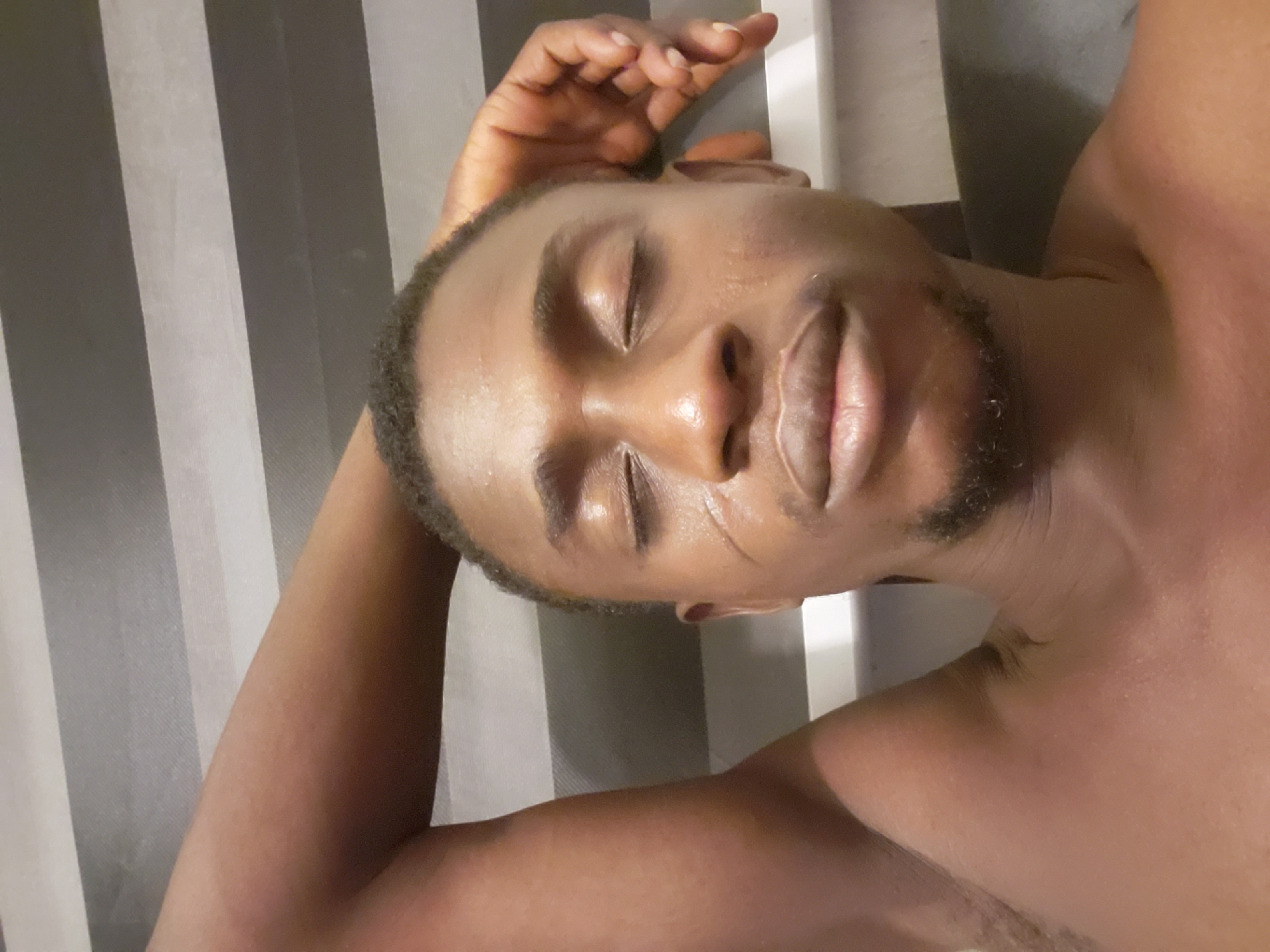The image presents a close-up, overhead view of an adult male of African descent, seemingly resting or asleep. He is shirtless, with his eyes closed and a calm expression. His skin has a visible sheen, which could indicate sweat or moisturizer. He has short, dark hair and stubble on his chin. His right arm is bent, with his hand positioned near his head. The background consists of distinct vertical stripes in varying shades of gray and white, suggesting window blinds, a curtain, or a patterned wall. The lighting appears soft and indirect, typical of an indoor environment. There is no visible text. The overall scene evokes a sense of peace or relaxation. The context places this scene in Adonkolo, Nigeria.