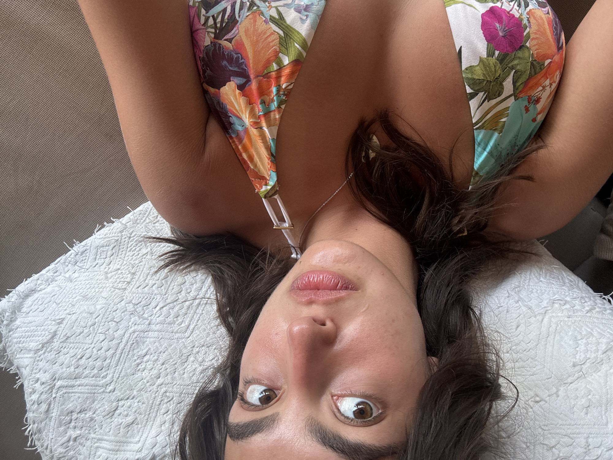 A woman with dark hair and brown eyes is lying on her back on a white, textured cushion or pillow. She is wearing a floral-print bikini top with a white strap and silver necklace. Her head is tilted, and her eyes are wide open, giving the impression of surprise or alertness. Her location is unknown, Mexico.