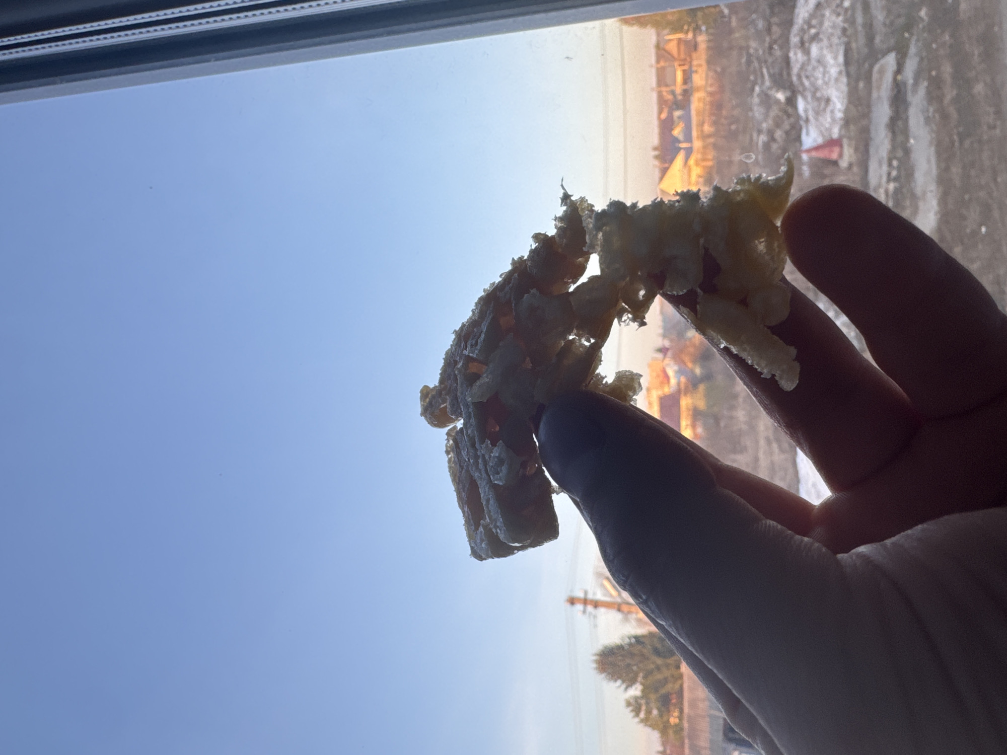 A rotated image depicts a person's hand holding a piece of light-colored, irregularly shaped food, possibly a fried pastry or dough, in silhouette against a bright outdoor scene. The hand and food are positioned centrally, casting dark shadows against the intense sunlight.
The top of the frame shows a dark window sill or frame. Beyond the hand and food, the background reveals a clear, blue sky. In the lower right portion of the background, through the window, a distant landscape is visible, appearing somewhat overexposed and blurred due to the bright light. This landscape includes what appear to be buildings or structures, and potentially power lines or utility poles. Patches of white on the ground, likely snow or ice, suggest a cold outdoor environment.
The bright sky and the warm, golden hue of the light in the distant background indicate that the photograph was taken during the daytime, possibly in the morning or late afternoon when the sun is lower in the sky. The activity captured is simply a person holding up food, perhaps for enjoyment or presentation, while looking out a window. The scene implies a view from indoors in Oktyabrsky, Russia, during clear, cold weather. No visible text is present in the image.