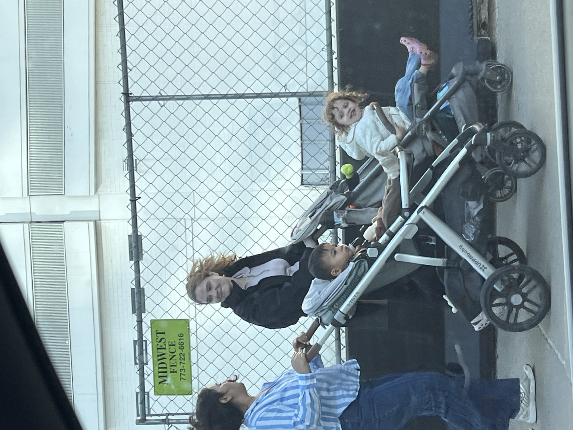 The scene depicts two adults with two children in strollers, all outdoors in Chicago, Illinois, United States. The adults appear to be interacting; one is wearing a blue and white striped shirt and jeans, looking up towards the other adult, who is wearing a black jacket over a light-colored shirt. The children are in Uppababy Vista strollers. One child is reclining in the stroller, while the other child is sitting upright. A chain link fence is visible in the background with a green sign that says "Midwest Fence 773-722-6616". Adjacent to the fence is a tall white building with horizontal grooves.