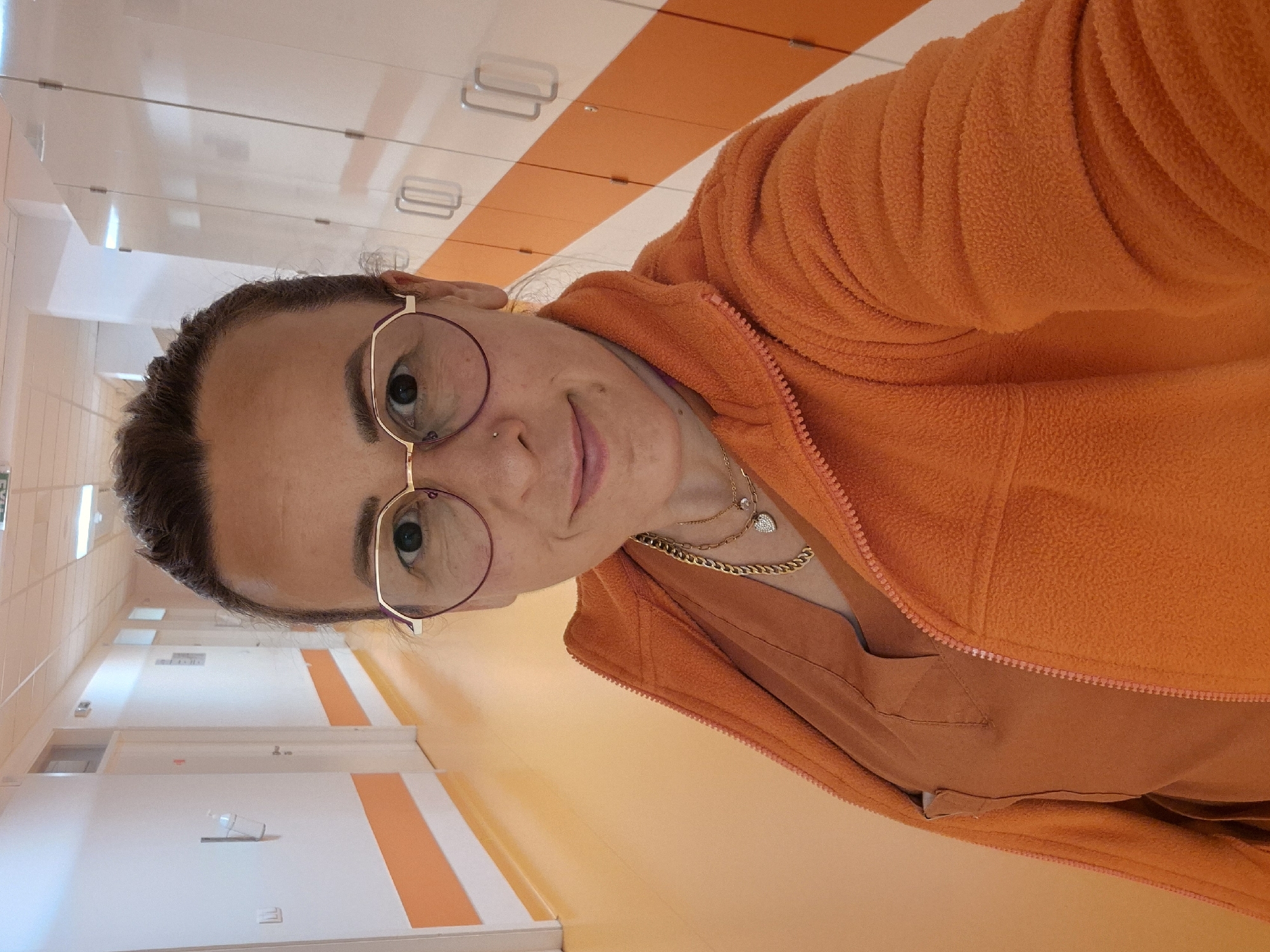 A woman with brown hair pulled back, wearing eyeglasses, a nose ring, a layered gold necklace, and an orange fleece jacket over a reddish-brown shirt, is facing the camera. The setting is an interior hallway with white walls featuring orange stripes near the floor. White doors are visible along the hallway. The location appears to be Unknown, Unknown.