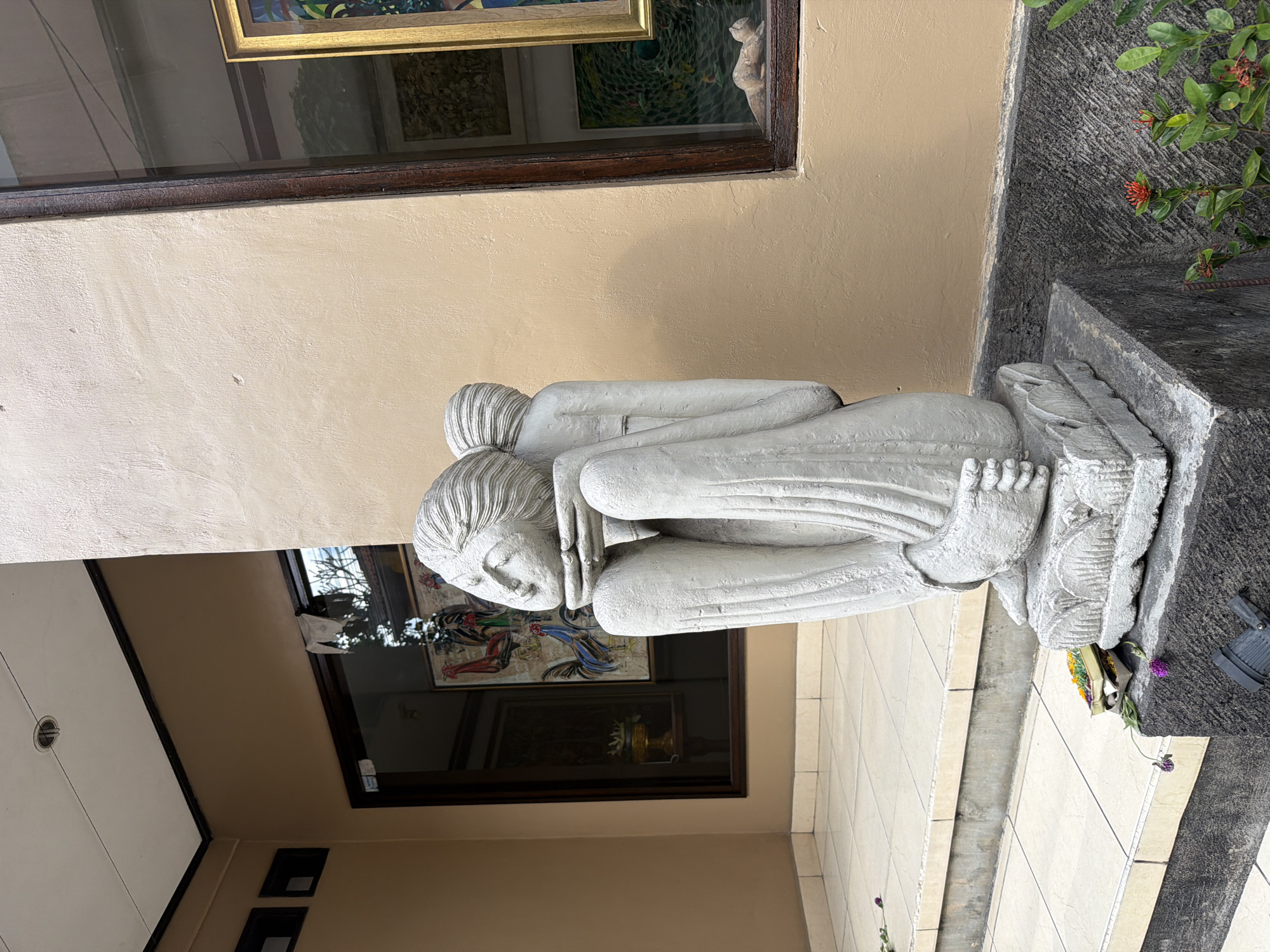 A stone sculpture of two intertwined figures, appearing to be in a seated, contemplative pose, is displayed outdoors in front of a building. The sculpture is light gray and detailed, with the figures' faces and draped clothing clearly defined. It rests on a dark gray stone pedestal.
Behind the sculpture, a building with a tan-colored facade features large windows. Through the windows, artwork, including paintings of roosters and a framed abstract piece, is visible. A small, light-colored animal figurine is placed on the windowsill of the right-hand window.
The foreground shows a tiled outdoor area with several steps leading up to the entrance. Small purple flowers and a small offering box filled with colorful petals are placed near the base of the sculpture and on the steps. Red ixora flowers with green leaves are growing in the lower right corner of the frame. The overall scene suggests an outdoor display, possibly at an art gallery or a cultural site in Ubud District, Indonesia, given the style of the artwork and offerings.
