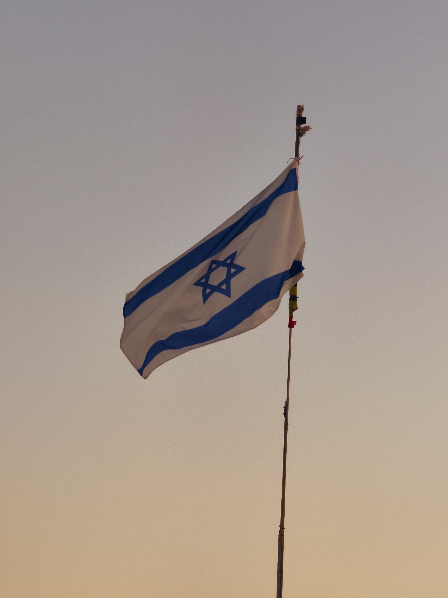 A flag of Israel, displaying a white background with two horizontal blue stripes and a blue Star of David in the center, is affixed to a thin, tall pole. The flag is partially unfurled and positioned in the upper left quadrant of the image. The pole, appearing to be made of bamboo or a similar material, extends from the bottom edge upwards, with some small red and yellow tags or ribbons near its top. The background consists of a gradient sky, transitioning from a pale gray at the top to a warmer, peachy hue towards the bottom, indicative of either dawn or dusk. The scene is devoid of any human figures or other visible objects in the immediate vicinity. The image appears to be captured outdoors, with no discernible urban or natural landscape elements beyond the sky. The location is identified as Lachish Regional Council, Israel.