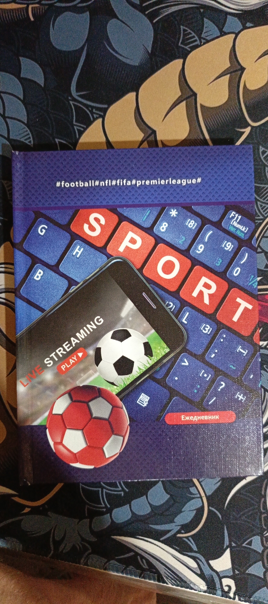 The image shows a close-up of a notebook or book cover. The cover has a dark blue textured background with a graphic design of stylized blue and black shapes. Dominating the center of the cover is a graphic of a smartphone screen displaying "LIVE STREAMING" with a "PLAY" button, and two soccer balls, one white and black and the other red and white. Behind the phone and balls is a graphic of a blue keyboard with large, red, white-lettered keys spelling out "SPORT". Above the keyboard graphic, there is text that reads "#football#nfl#fifa#premierleague#". At the bottom of the cover, there is a red rectangular button with white Russian text that reads "Ежедневник", which translates to "Diary" or "Notebook". The notebook is lying flat, with its spine facing down and slightly to the right. The background outside the notebook appears to be a surface with a similar dark blue and black pattern. There is no identifiable location in the city of Kalininskaya, Russia.