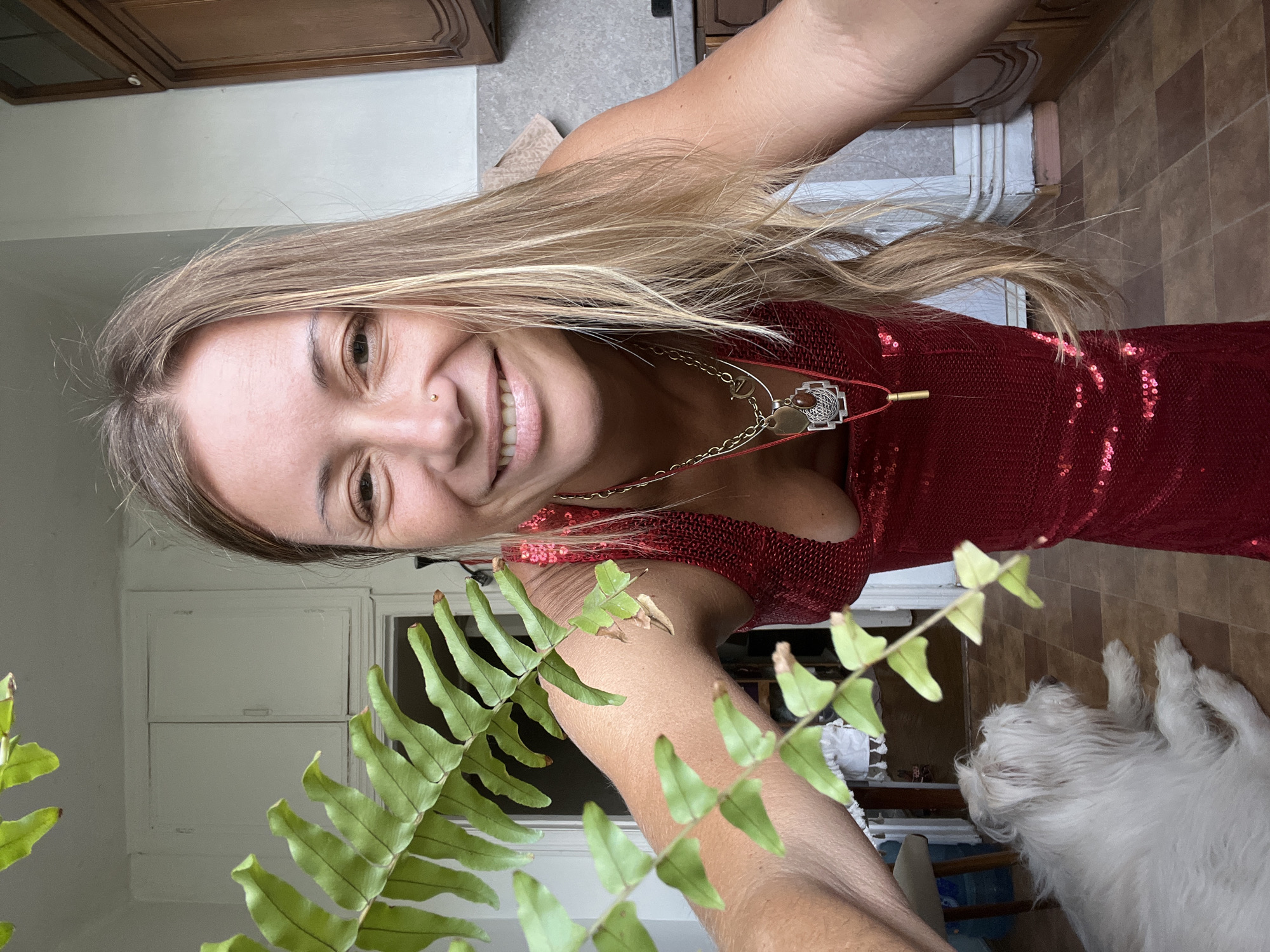 This is a close-up, selfie-style photograph of a woman wearing a red sequined dress and a necklace. She has long, blonde hair and is smiling at the camera, with a nose ring. In the foreground, a green fern plant partially obscures the view. In the background, a white, fluffy dog is visible, lying down. The setting appears to be indoors, possibly a kitchen or dining area, with wooden cabinets and tiled flooring. The lighting suggests it is daytime. The woman's expression is happy and relaxed. There is no visible text in the image.