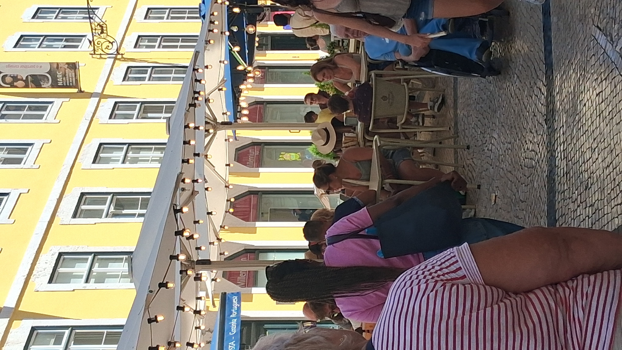 This outdoor scene captures a bustling European cafe setting in Lisbon, Portugal, likely in the Baixa district given the characteristic yellow building with white trim. The facade of the building is painted a vibrant yellow and features multiple windows with white frames. Strings of bare lightbulbs are draped over cafe umbrellas, suggesting it is either late afternoon or early evening.
Several tables are occupied by patrons enjoying drinks and conversation. In the foreground, on the left, a person wearing a pink and white striped shirt is partially visible, facing away from the camera. To their right, a woman with long, dark braided hair, wearing a pink shirt and carrying a dark blue bag, is seated. Further back, a woman in a green tank top and denim shorts is seated at a table, wearing sunglasses and a straw hat. Another woman with blonde hair is seated across from her.
In the background, a person in a wheelchair, wearing blue pants, is visible. Other individuals are seated at various tables, engaged in typical cafe activities. The ground is paved with traditional Portuguese cobblestones, arranged in a mosaic pattern. The atmosphere appears lively and relaxed, indicative of a popular dining and social spot in the city.