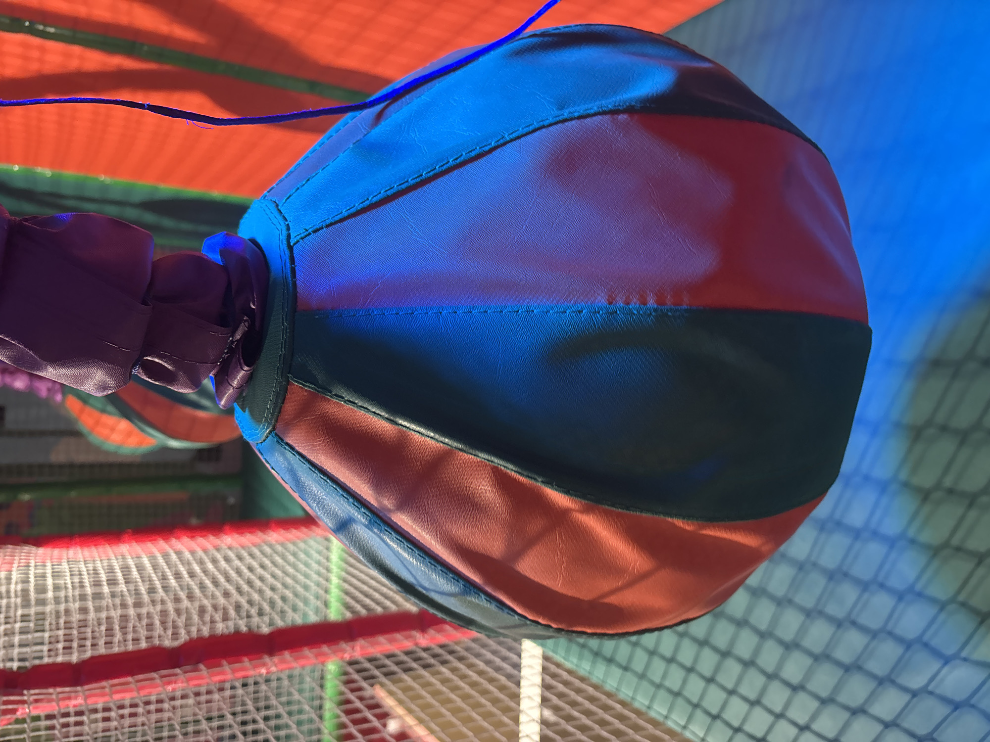 A close-up, low-angle shot focuses on a hanging, multi-colored punching bag within an indoor play area. The punching bag is segmented into sections of red, dark green, and blue, with visible stitching outlining the panels. A purple fabric loop attaches the punching bag to an unseen support above.
The background is a blur of colors and textures suggestive of a soft play or adventure zone. To the left, a white net enclosure with red framing is visible, partially obscuring the view. The right side of the background features vibrant orange and green vertical stripes, illuminated by blue and red lighting that casts reflections on the punching bag and the net. The floor appears to be a light blue surface, with the net casting a grid-like shadow. The overall scene suggests an active, possibly entertainment-oriented environment. The image does not provide specific details about its location within Dubai, United Arab Emirates.
