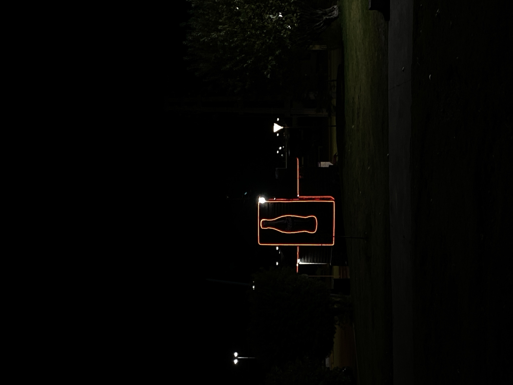 At night, an illuminated orange neon Coca-Cola sign stands vertically, framed by a square, with an adjacent vertical line, atop a pole. The sign features a bottle silhouette. It is located in Ushuaia, Argentina. Dim streetlights can be seen. Behind the sign is a dark structure, with trees and grass to the right of the sign. The background is predominantly black.