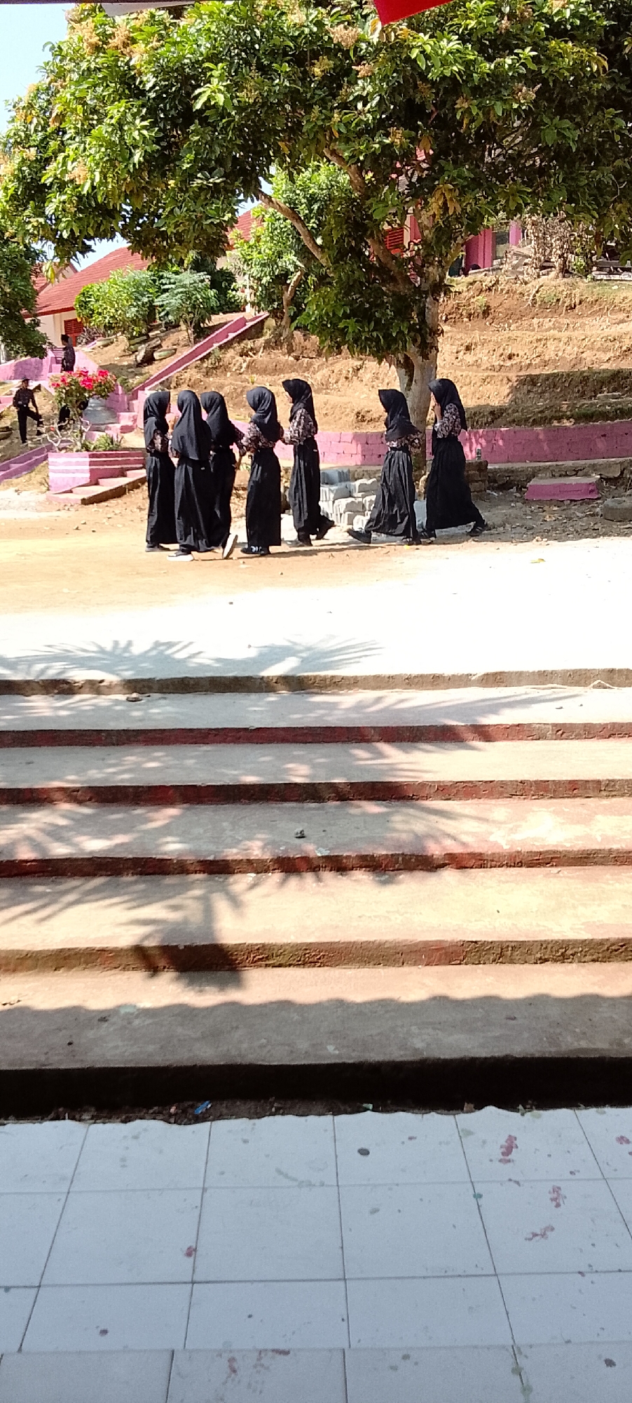 A group of six young girls wearing black hijabs and black dresses are walking in a line on a dirt path. The girls are positioned in the middle ground of the image. Behind them, there are buildings with red roofs and a large tree with green foliage. In the foreground, there are concrete steps with red trim. The overall environment appears to be an outdoor school or religious institution setting. The image was captured in Jepara, Indonesia.