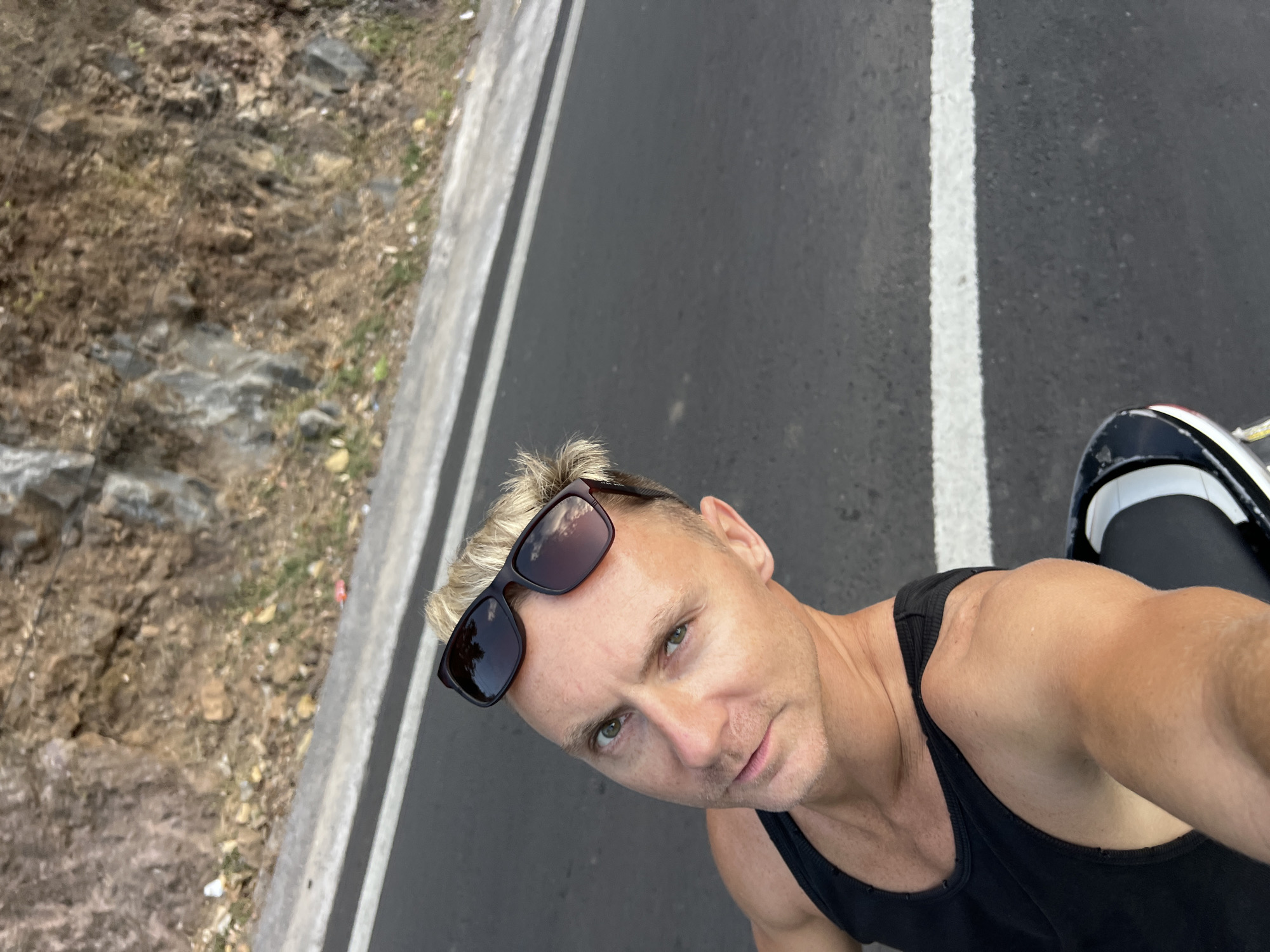 An outdoor, high-angle, medium shot captures a fair-skinned man with blond hair and green eyes, wearing a black tank top and sunglasses, sitting on what appears to be a motorcycle or scooter. He is looking towards the camera, with his right arm visible. The man is positioned in the foreground, against a backdrop of a paved road with a white line, a rocky, dry hillside, and sparse vegetation. The scene is brightly lit, suggesting daytime. The specific location is Abang, Indonesia.