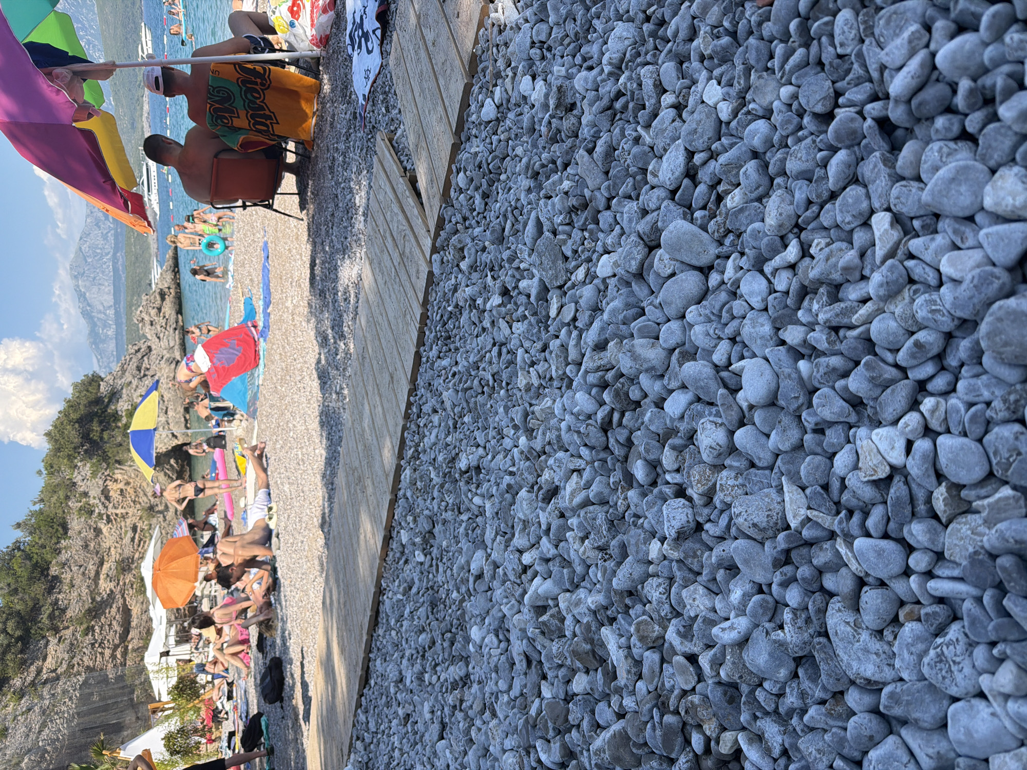 The image depicts a crowded beach scene in Radovići, Montenegro, under a clear, sunny sky. The foreground is dominated by a dense collection of grey pebbles. In the midground, several beachgoers are relaxing on towels and chairs. Some are seated under a multi-colored umbrella, with two men in chairs and another man lying on a towel in front of them. Further back, people are wading in the shallow, blue water, with some children playing. The background features a rocky cliff face dotted with trees, and beyond that, a body of water with boats and distant mountains. Several umbrellas of various colors dot the beach, providing shade. The overall impression is a lively summer day at the beach.