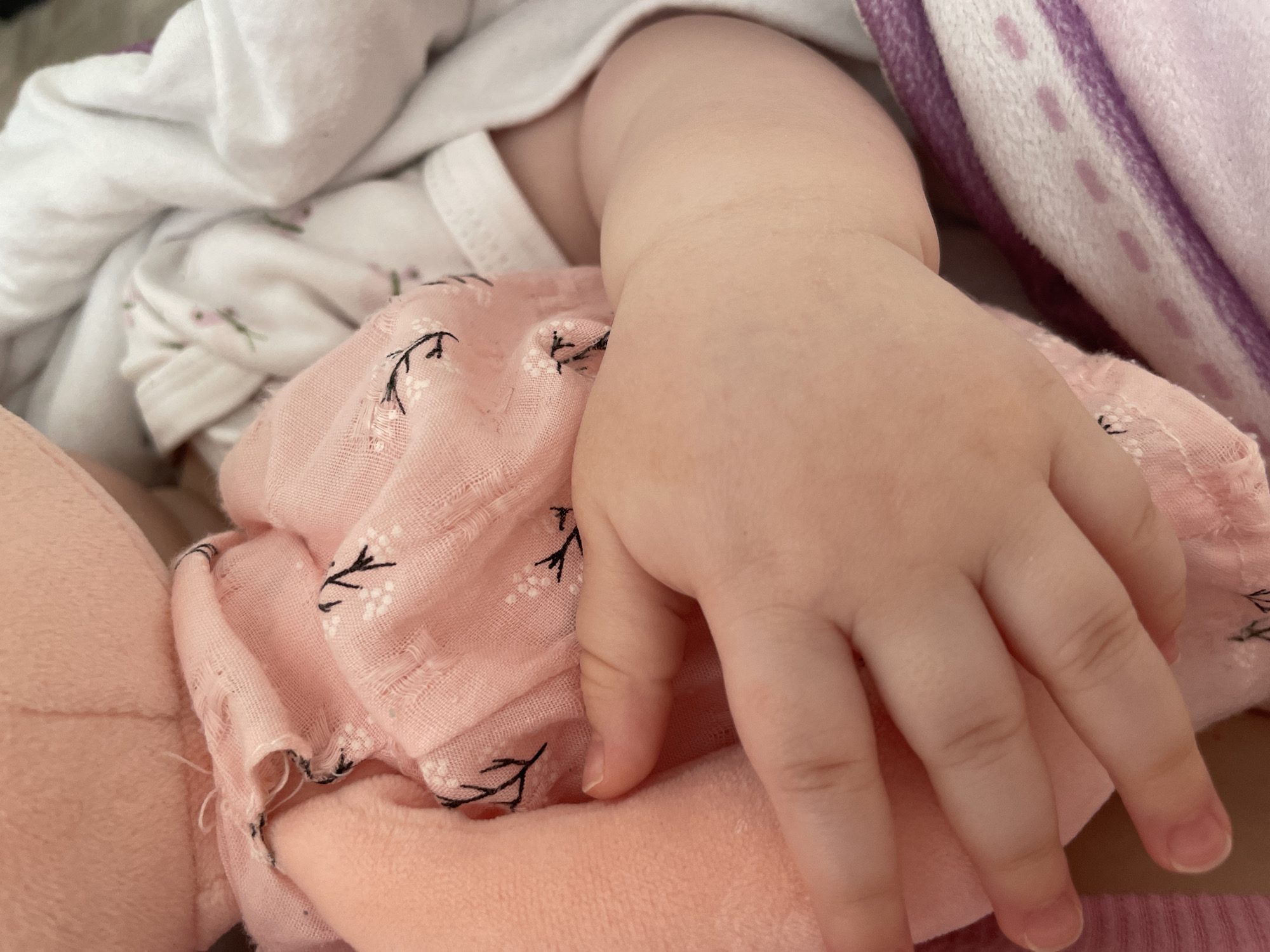 A close-up photograph depicts a baby's arm and hand resting on soft fabric, possibly a swaddling blanket or doll. The baby is wearing a white onesie with a floral pattern and is partially covered by a pink floral patterned cloth. The baby's hand is pink and chubby. The photograph does not provide enough information to infer any location, including Chaikovsky, Russia.