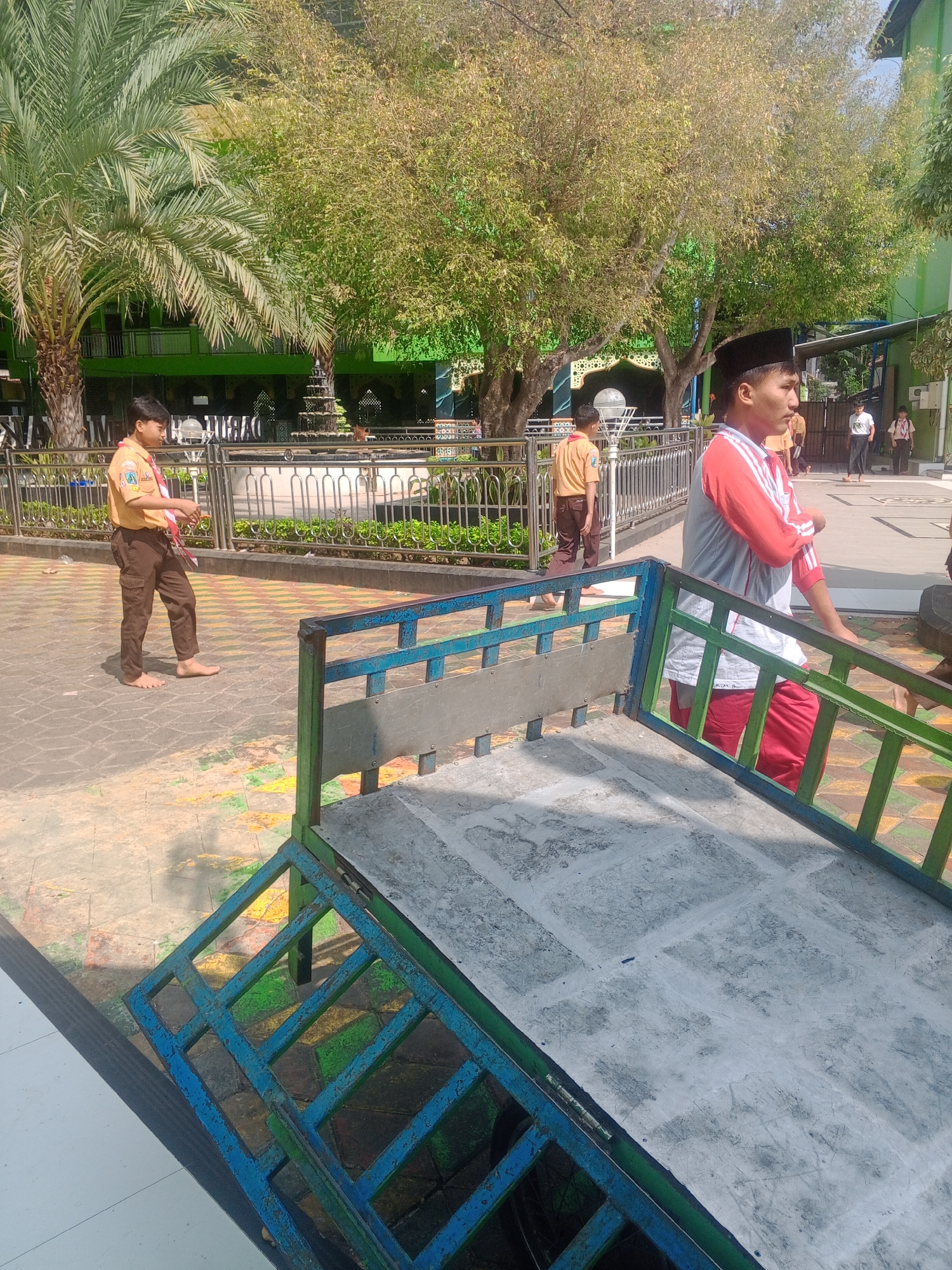 The image depicts a scene outdoors, possibly in a school or mosque complex in Ponorogo, Indonesia. In the foreground, a metal structure with blue and green painted bars and a concrete surface is visible. A young person wearing a red and white shirt and red pants stands to the right, facing away from the camera, with their profile visible. Behind them, another young person in a scout uniform of a tan shirt and brown pants is walking barefoot on a patterned brick pathway. Further in the background, more individuals, also in school or religious attire, are seen walking. Trees, one with large fronds resembling a palm tree, are prominent in the background, along with a green building and a fountain. The overall environment suggests a sunny day in an urban or semi-urban setting.