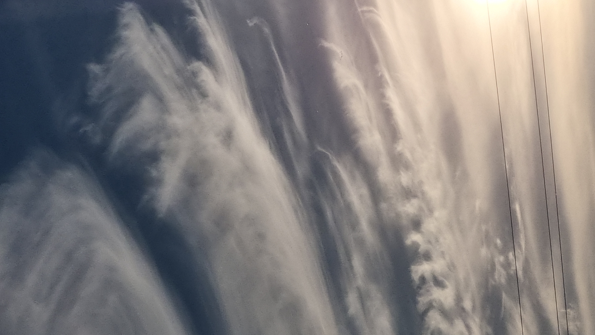 The image displays a dramatic sky filled with wispy, streaky clouds that appear to be blowing horizontally. The clouds transition from a dark, deep blue on the left side to a bright, sunlit expanse on the right, where the sun is partially visible, creating a luminous glow. Three thin, dark lines, likely power lines, cut vertically through the right side of the frame, extending upwards into the bright sky. The cloud formations suggest a windy day, possibly with the approach of a weather front. The overall mood is one of awe and the immense power of nature. The scene is set outdoors, likely in Vysokoe, Russia, given the context. The time of day appears to be either early morning or late afternoon, judging by the angle and intensity of the sunlight. There is no visible text in the image.