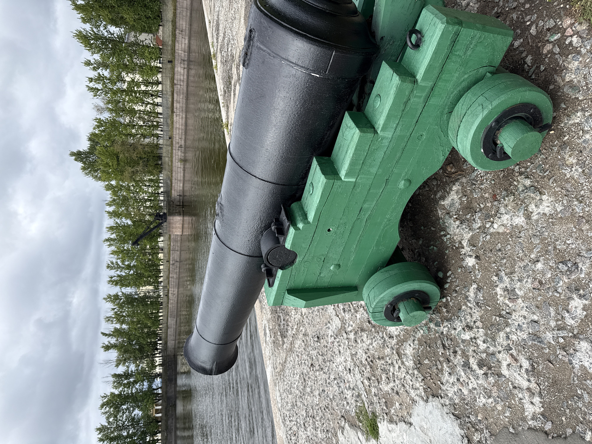 The image depicts an old black cannon with a green wooden carriage placed on a stone embankment next to a body of water. In the background, a line of trees separates the water from buildings and a tall industrial crane. The sky is overcast with grey clouds. The cannon is positioned in the foreground, angled slightly to the left, with its barrel extending towards the viewer. The wooden carriage has visible planks and wheels. The stone embankment shows signs of wear and tear, with scattered pebbles. The water appears calm and reflects the overcast sky. The overall scene suggests a historical or maritime location, possibly a park or museum area. The location is identified as Kronstadt, Russia.