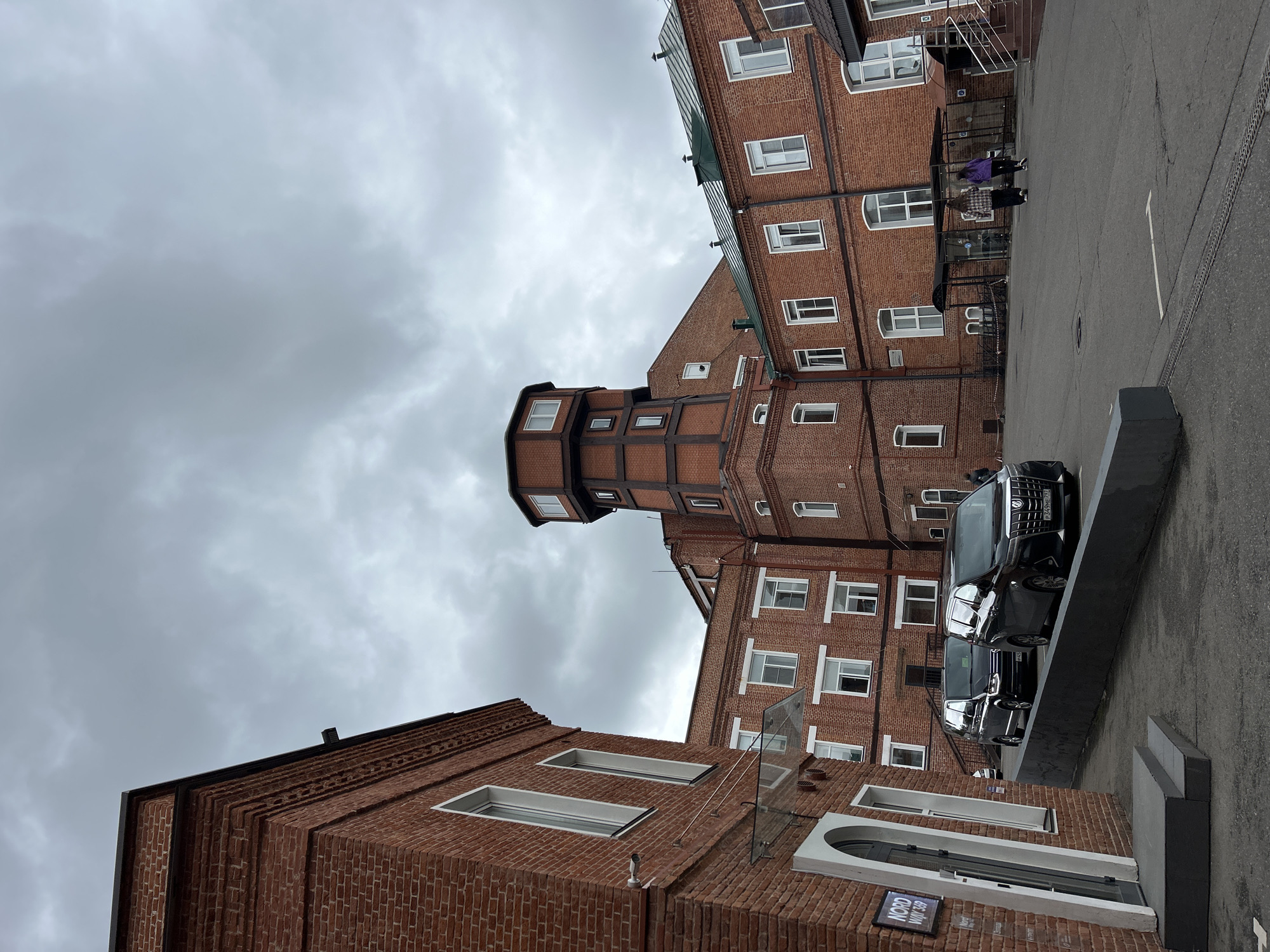 The image shows an exterior view of a brick building complex under an overcast sky. The architectural style is characterized by red brick facades, white-framed windows, and a distinctive tower-like structure with brown trim. Several modern vehicles are parked near the building, adjacent to a gray concrete structure. Two individuals are walking in the background near a covered walkway. A sign is visible near a white-framed entrance at the bottom left. The location is Unknown, Unknown.
