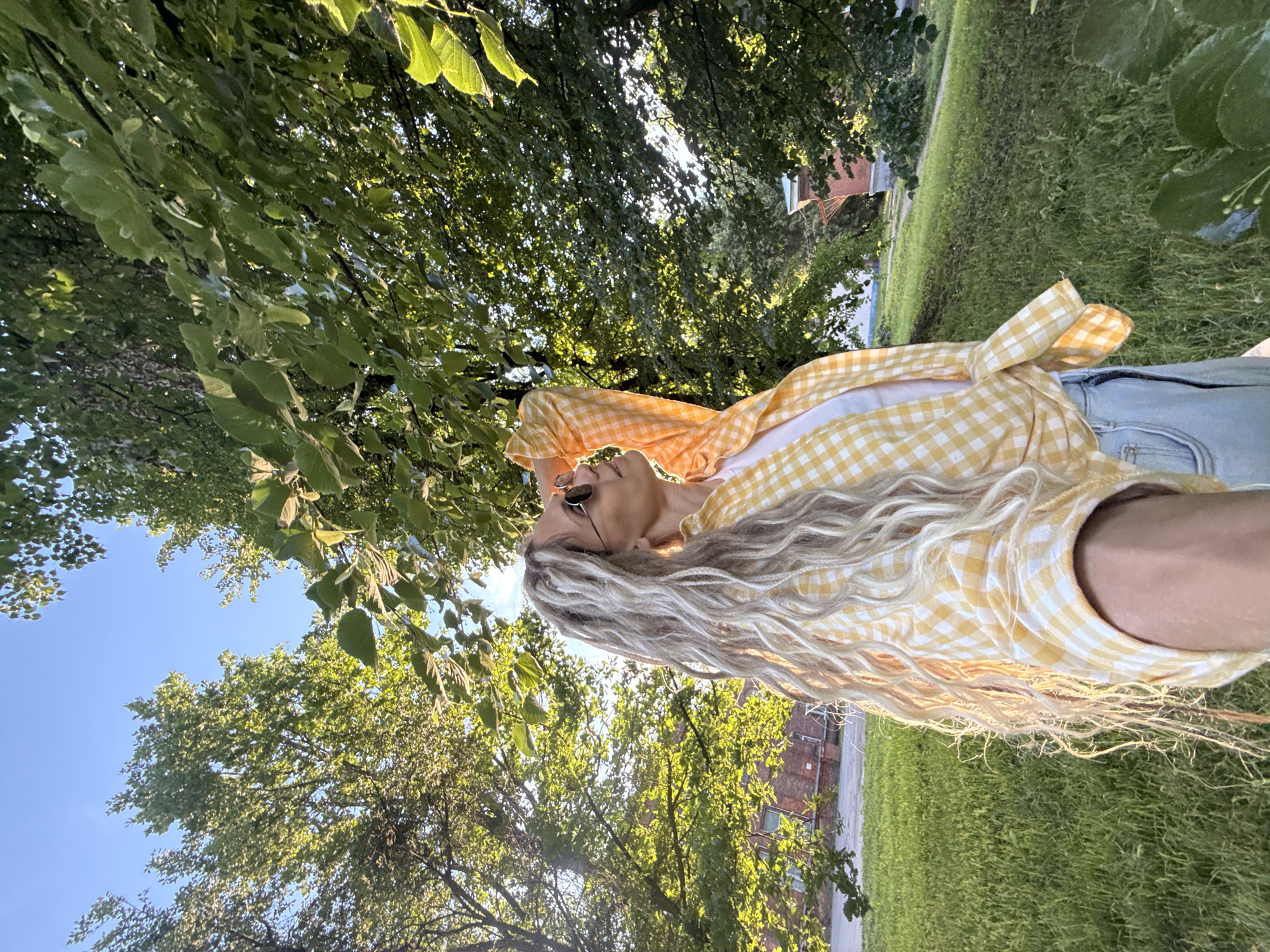 A woman with long, blonde dreadlocks stands in a park under a canopy of green trees. She is wearing a yellow and white checkered shirt over a white t-shirt and light blue jeans. Her hand is raised, resting on the back of her head, and she is looking up and to her right. Round sunglasses obscure her eyes. The sun is shining through the leaves, creating dappled light on the grass and the woman. In the background, a brick building with white trim is visible. The ground is covered in lush green grass, and the sky is a clear blue. The location is identified as Shebunino, Russia.
