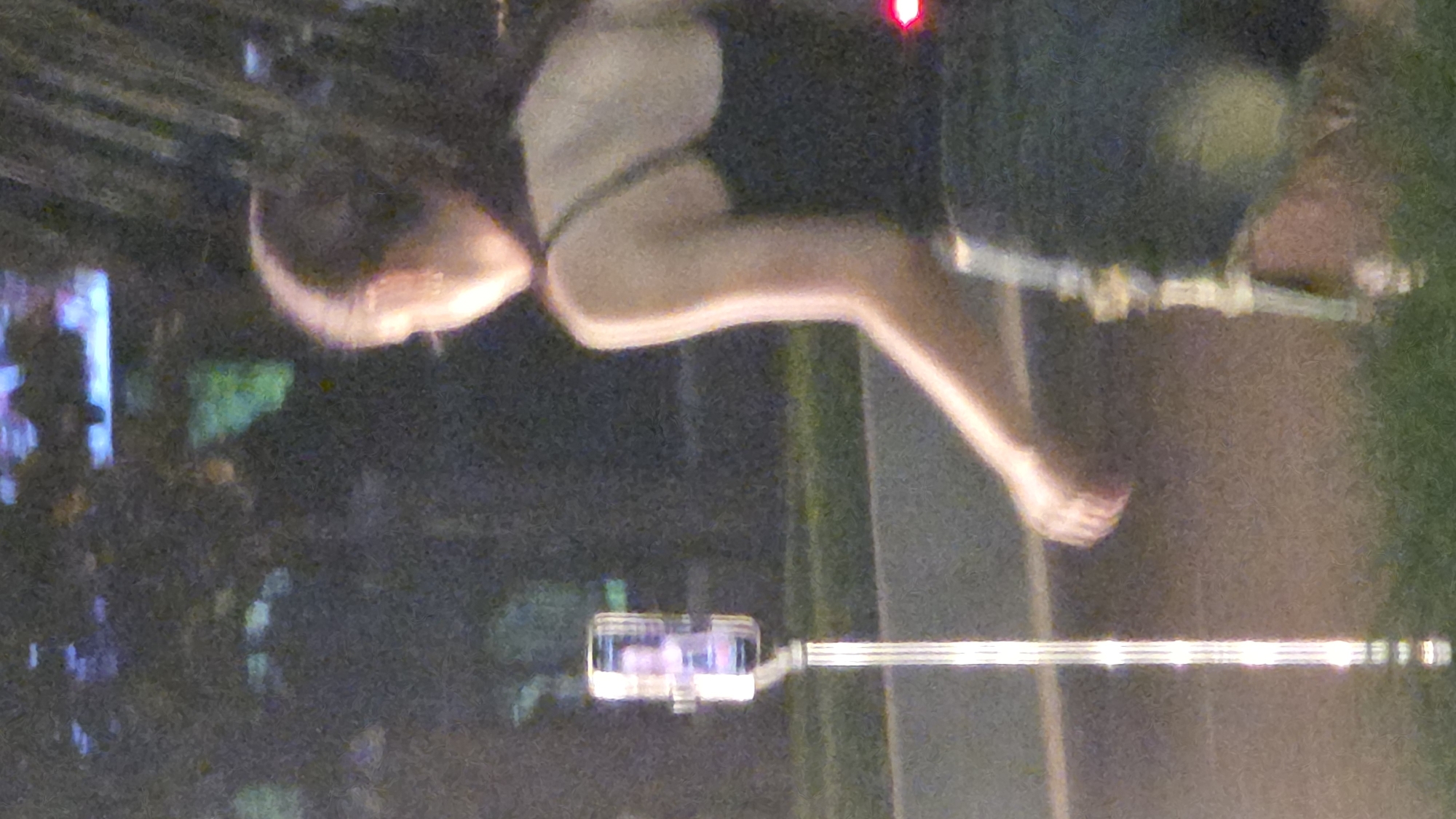 The image depicts a female adult viewed from the side. She is wearing a dark-colored top and is barefoot. She appears to be positioned on a horizontal pole or bar, possibly performing some form of exercise or activity. The background is blurred and indistinct but includes possible building lights. The location is indicated to be Nha Trang, Vietnam, but there are no distinguishable landmarks to confirm that specific location from the image alone. A cable runs across the bottom of the image.