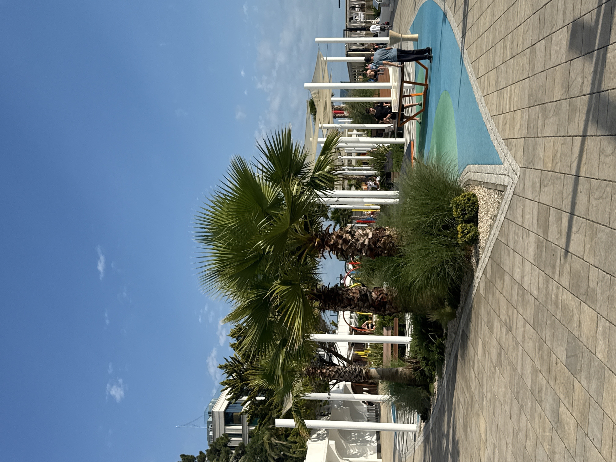The image captures a sunny day in Sochi, Russia, likely at a public recreational area. In the foreground, a paved walkway made of rectangular stone tiles leads into a vibrant scene with palm trees and lush greenery. A portion of the walkway transitions into a colorful, curved surface painted with blue and green sections, suggesting a play or activity area.
Several palm trees stand prominently, their fronds reaching towards the clear blue sky dotted with a few wispy clouds. Behind them, a modern building with white walls and balconies can be seen, surrounded by trees. In the mid-ground, a ping pong table is set up on the blue and green surface, with two young men actively engaged in a game. One person, wearing a dark t-shirt, is positioned behind the table, while the other, in a blue t-shirt and dark pants, stands facing him.
Further back, several white columns support canopies, providing shade over seating areas where other people are visible. A playground structure with colorful elements is also partially visible in the distance. The overall atmosphere is one of leisure and outdoor activity, characteristic of a resort town like Sochi.