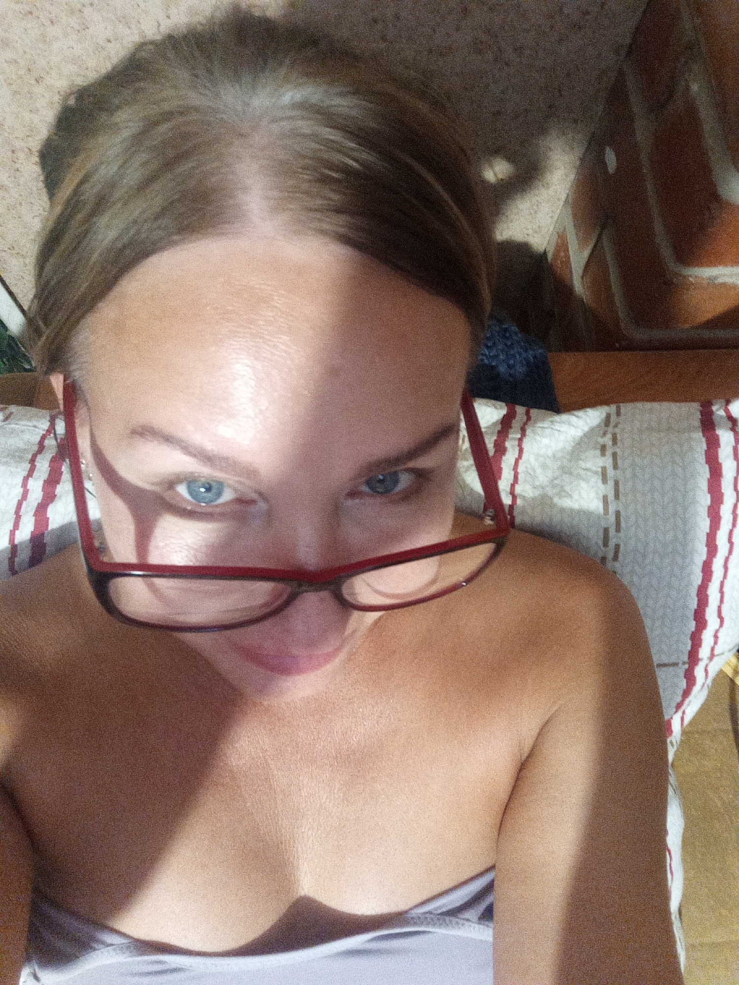 An overhead, close-up, indoor selfie shows a woman with light brown hair and blue eyes wearing red-framed glasses. She is looking upwards towards the camera with a neutral expression. Her shoulders and upper chest are visible, and she appears to be lying down on a pillow with red and white stripes. The background includes what looks like a brick wall and a textured surface. The lighting suggests it is daytime, with shadows cast across her face and body. The image was taken in Novopokrovka, Russia.