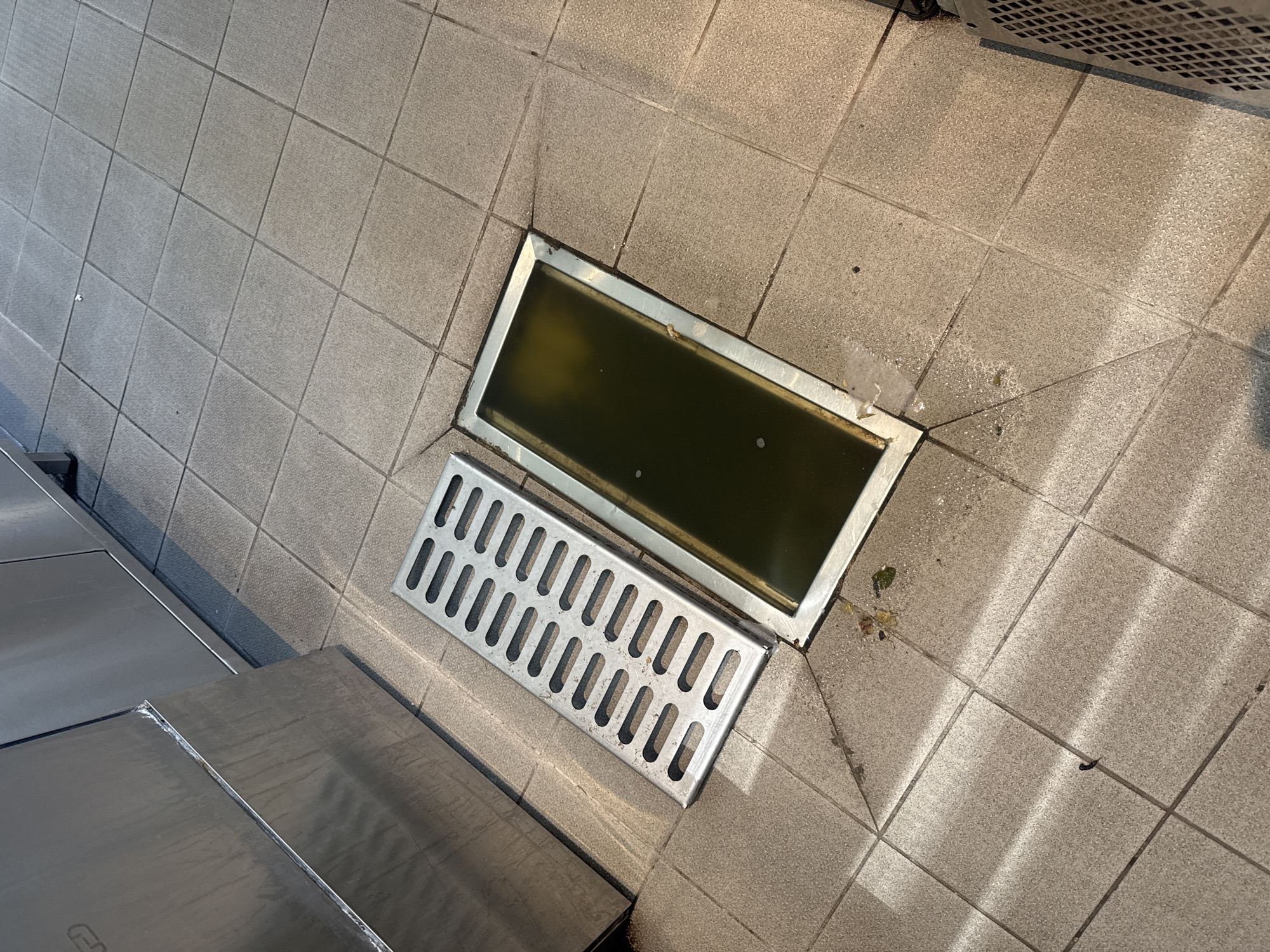 The image shows a tiled floor with a rectangular metal drain filled with dark, likely stagnant, water. The drain cover, a separate metal grate, is placed next to the drain. Part of a stainless-steel structure is visible on the left side of the frame. The location is Tel-Aviv, Israel.