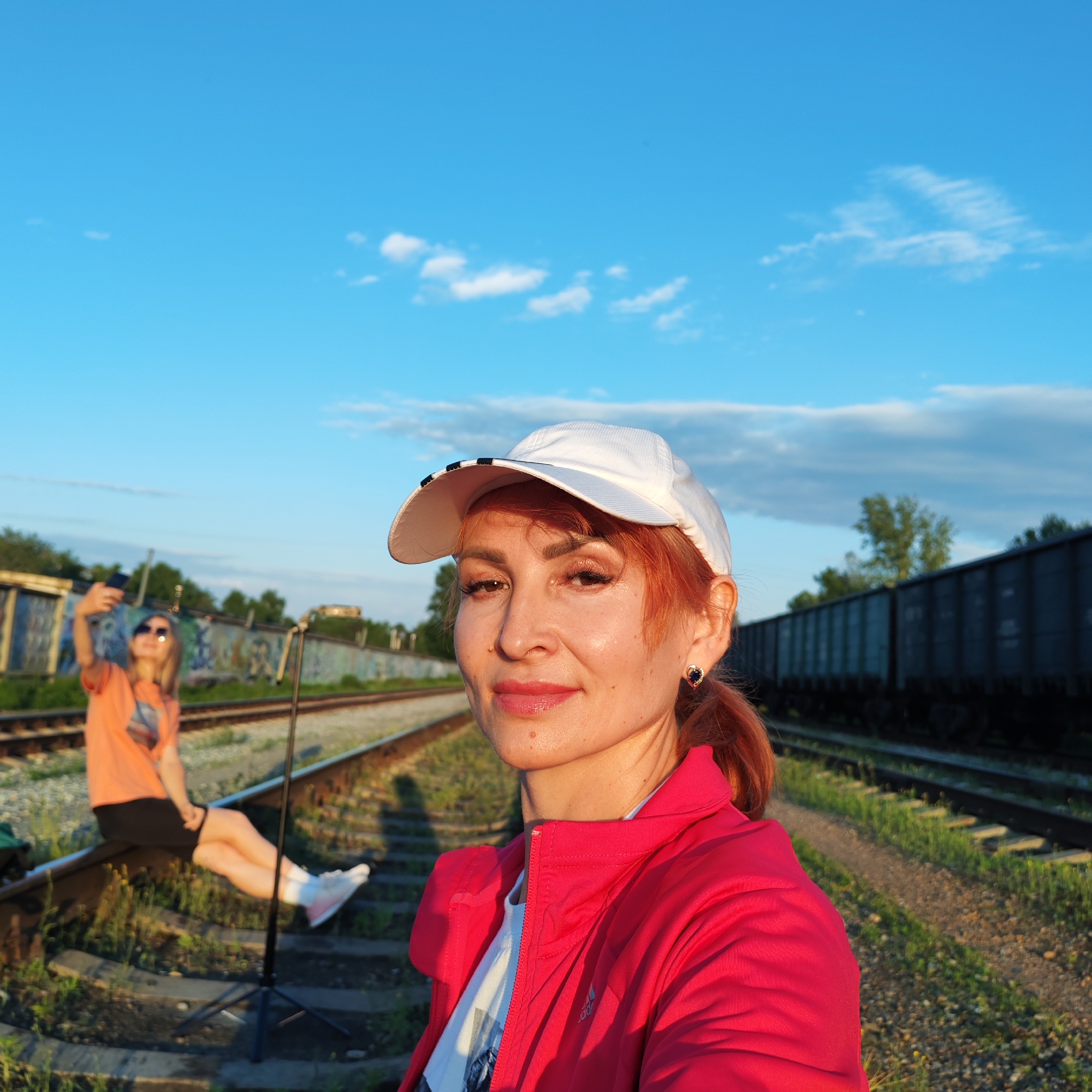 A selfie captures a woman with red hair, wearing a white baseball cap and a bright pink jacket, posing on a railway track under a clear blue sky with scattered clouds. Her face is turned towards the camera, and she has makeup on, including lipstick and eyeshadow. A young woman with blonde hair and sunglasses, wearing an orange t-shirt and black shorts, is visible in the background, sitting on the railway track and holding up a smartphone, seemingly taking a photo or video of herself. She is wearing white sneakers. A black tripod stands between the two women. In the background, a long train composed of dark freight cars is parked on a parallel track. The environment suggests an outdoor, possibly industrial or semi-abandoned location. The railway tracks run through an area with some vegetation and graffiti on a distant structure. The lighting indicates it is either early morning or late afternoon, with the sun casting long shadows. The city of Chaikovsky, Russia, is known for its industrial facilities, including a large chemical plant, and its proximity to the Kama River, which could explain the presence of railway infrastructure and freight trains.