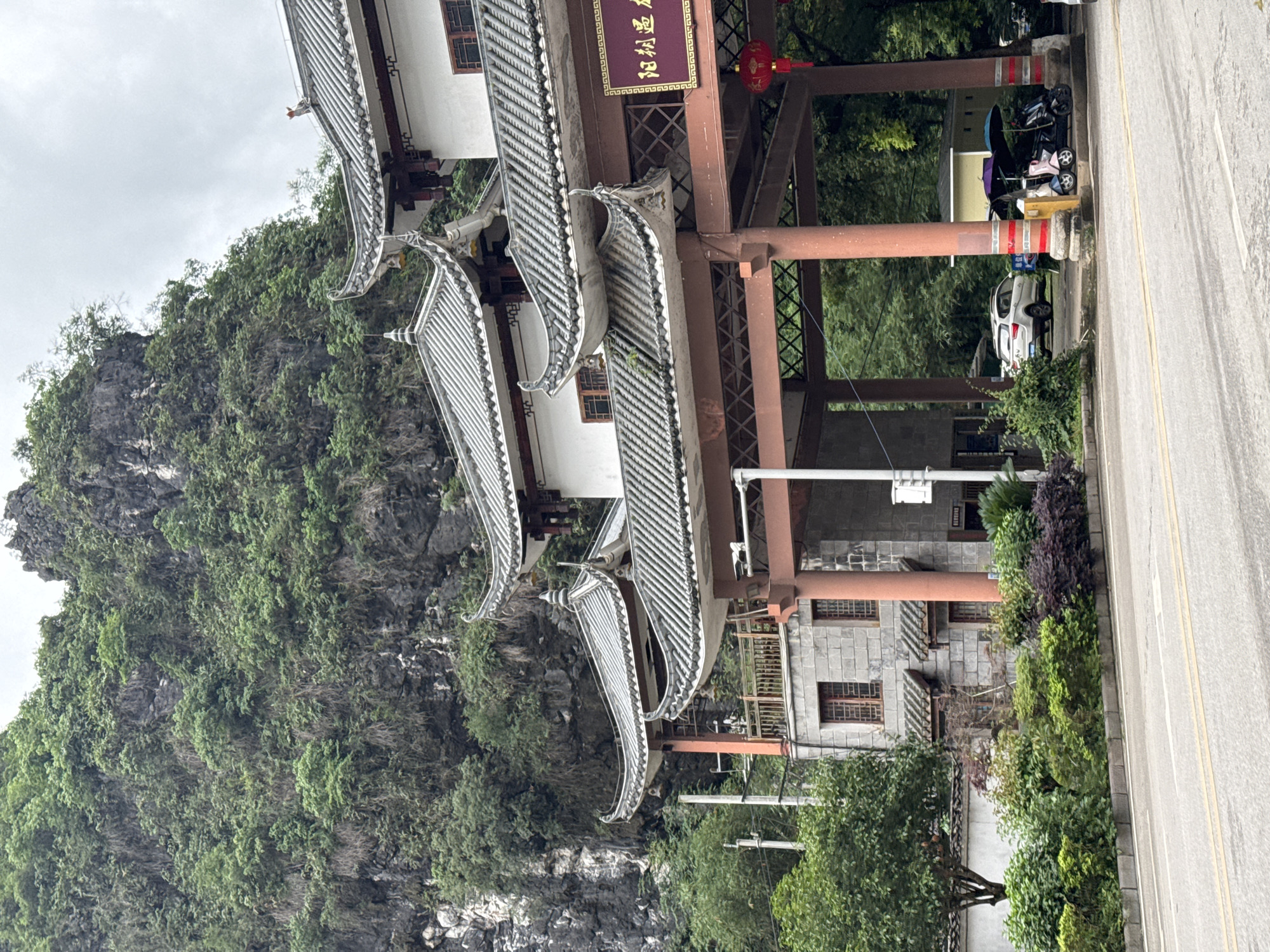 The image shows an Asian-style structure with multi-tiered, gray tiled roofs situated next to a mountain covered in dense green vegetation. The structure has reddish-brown support beams and a sign with gold characters, suggesting a cultural or historical significance. To the right, a road is visible, and parked vehicles including a white car and a stroller can be seen. The photograph was taken during the day with overcast skies, giving the scene a somewhat muted appearance. The location is implied to be in or near Guilin, China, due to the traditional architecture and the karstic landscape featuring green hills and mountains.