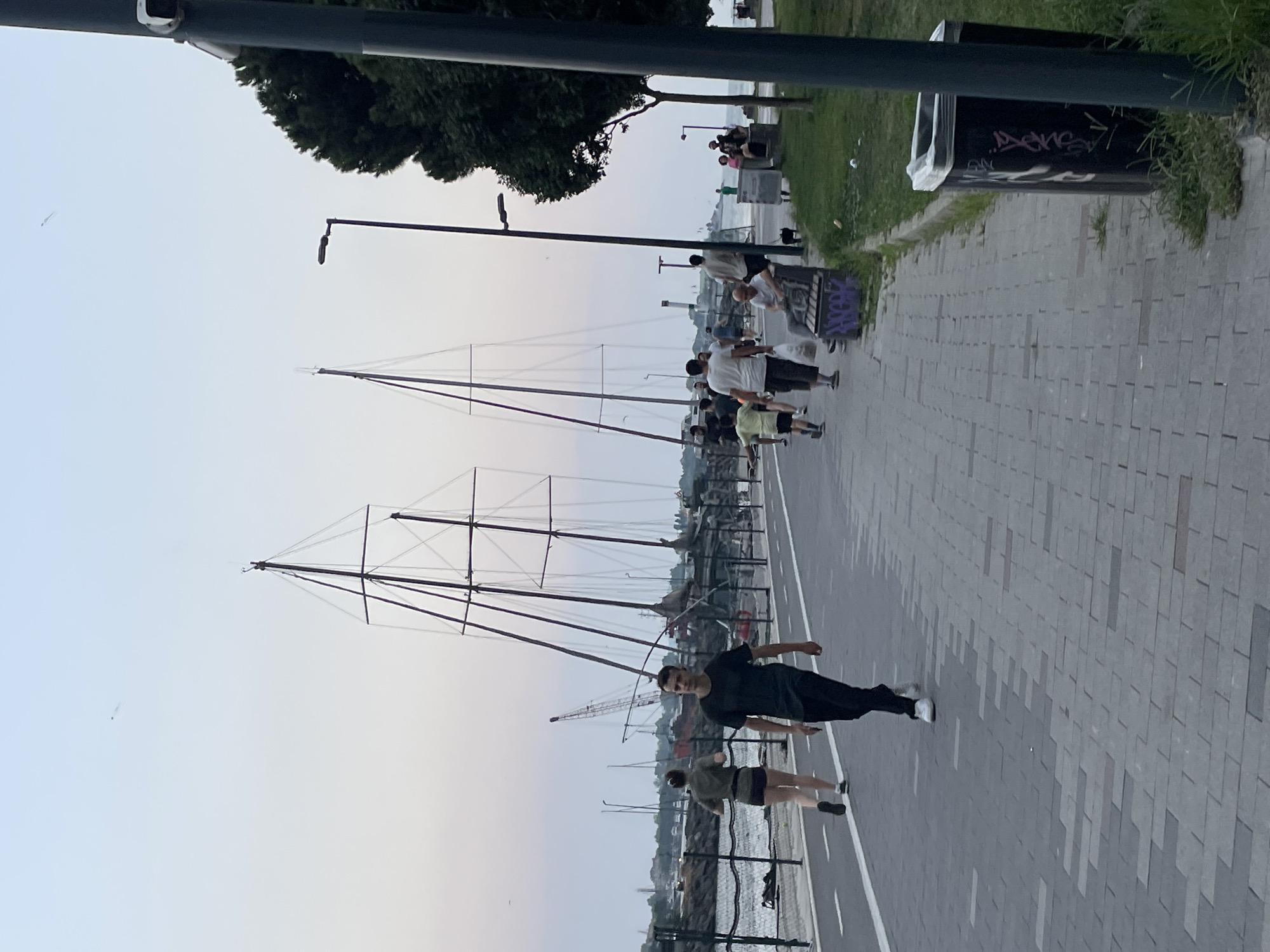 An outdoor, eye-level, full shot captures a promenade near the sea in Kadıköy, Turkey. The image is dominated by the sky, which is a pale gradient of blue and pink, suggesting dusk or dawn. In the background, two large, skeletal boat masts with intricate rigging rise prominently against the sky, indicating the presence of a marina or boatyard. The promenade itself is paved with grey bricks laid in a patterned manner.
Several individuals are present on the promenade. In the foreground, a young man with short dark hair, wearing a black t-shirt and black pants with white sneakers, walks towards the viewer. Slightly behind him and to his left, a woman with dark hair pulled back, wearing a khaki t-shirt and dark shorts with white sneakers, is jogging away from the viewer.
Further down the promenade, a group of people are visible. To the left of the boat masts, a crane is partially visible, suggesting ongoing construction or maintenance. On the right side of the promenade, a dark green tree with dense foliage stands tall. A dark grey trash can, graffitied with white and pink markings, is positioned near the tree and the edge of the promenade. Several people are seated on benches to the right, some appearing to be resting or conversing. The sea is visible on the left, separated from the promenade by a black metal fence. The overall atmosphere suggests a leisurely scene in a coastal urban environment.