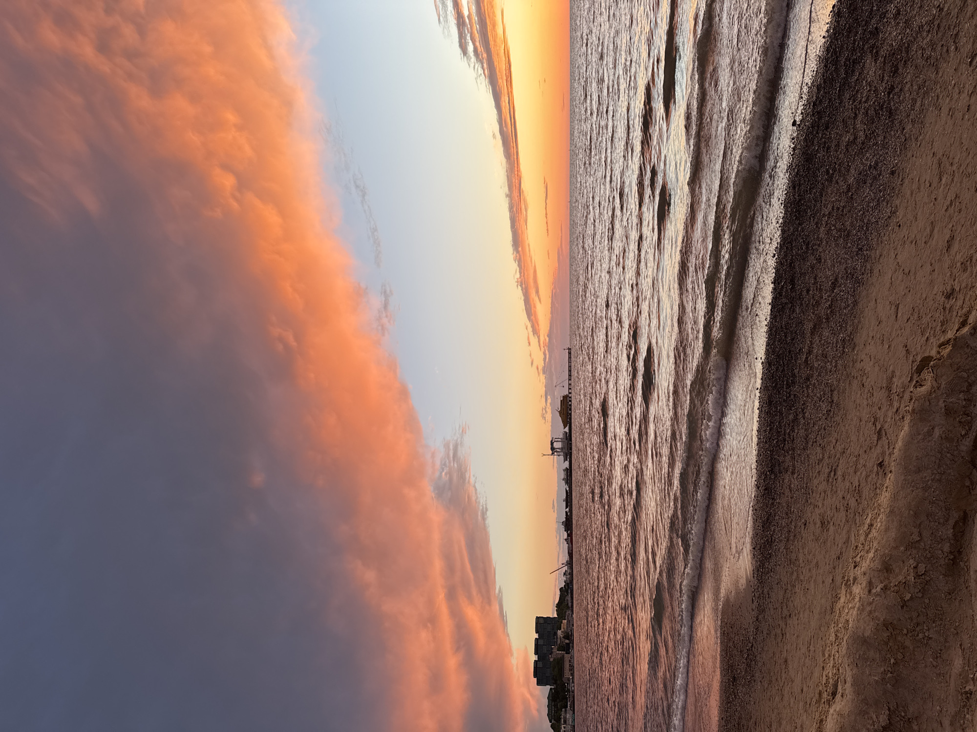 The image displays a vibrant sunset or sunrise over a coastal scene, likely in Anapa, Russia. The sky dominates the left side of the frame with a dramatic display of clouds. Large, fiery orange and pink clouds are prominently featured, catching the warm light, while a darker, purplish-grey cloud mass sits beneath them on the far left. The colors transition from intense orange to softer hues and pale blue further to the right.
The horizon line extends across the middle, separating the colorful sky from the sea. The water, occupying the right side of the image, is textured with numerous small waves, reflecting the ambient light with shades of muted brown, orange, and grey.
In the foreground, a sandy beach stretches across the bottom, appearing dark and textured, meeting the water's edge. Along the distant horizon, silhouettes of buildings, possibly part of a city skyline, and industrial structures, such as cranes or piers, are visible against the glowing sky. There are no people visible, and the overall activity is limited to the natural event of the sun rising or setting over the coast. The scene evokes a sense of dramatic serenity and natural beauty at a specific time of day characterized by warm, low-angle light. No visible text is present.