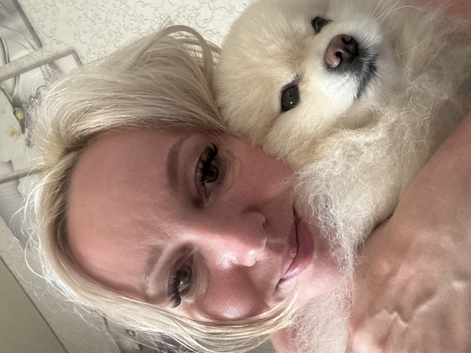 A close-up selfie captures a woman with blonde hair and visible eyelashes holding a fluffy cream-colored Pomeranian dog. The woman's face is turned towards the camera, with her left cheek pressed against the dog. Her lips are slightly parted, and she appears to be smiling. The dog's head is resting on her shoulder, with its eyes looking towards the camera. The background is blurred, showing a textured wallpaper with a light pattern. The image is taken indoors, likely in a residential setting. The specific location in the city of Komsomolsk-on-Amur, Russia, cannot be determined from the visual information provided in the image.