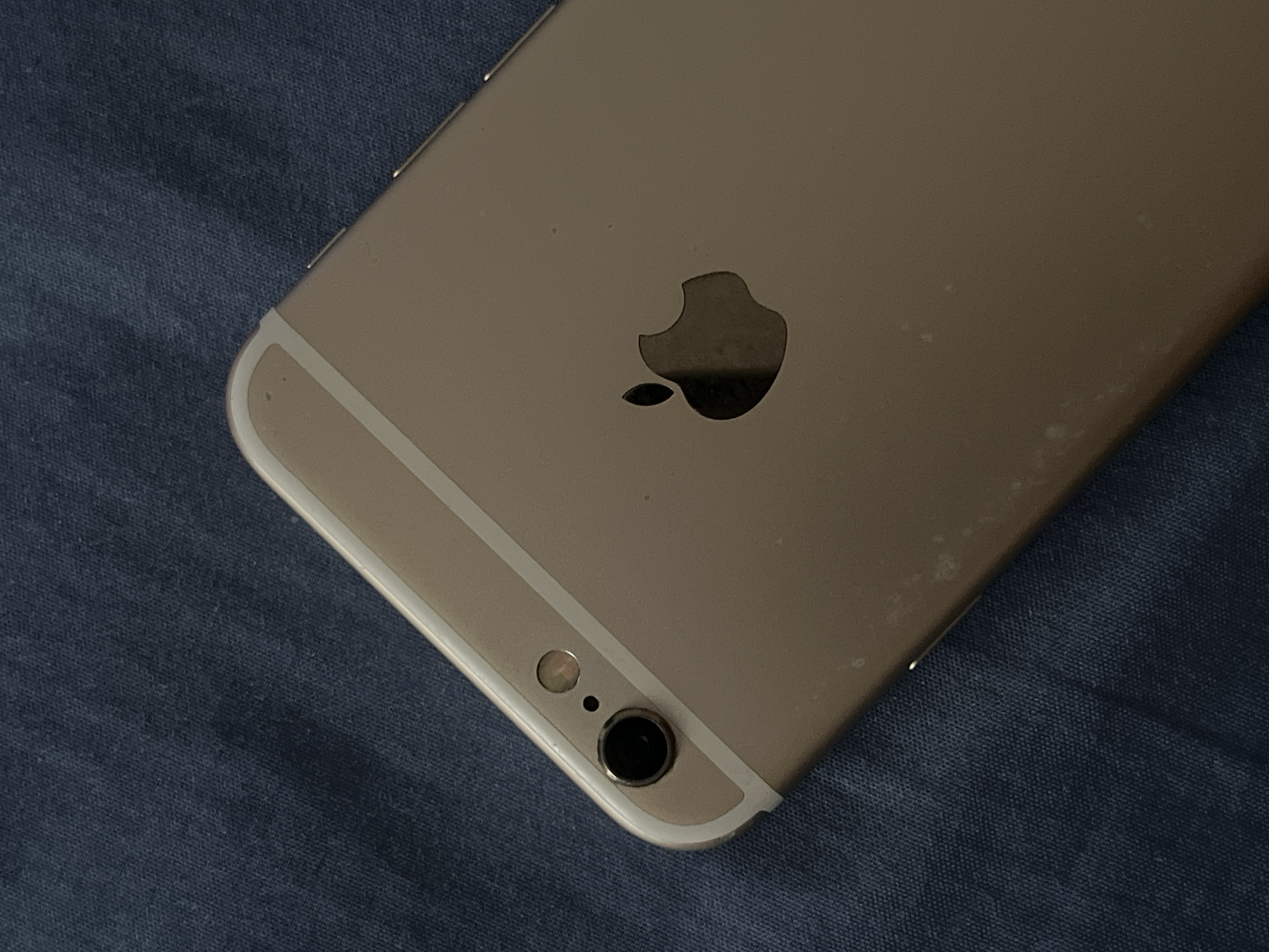 A close-up, overhead shot shows the back of a rose gold iPhone 6 Plus. The phone is resting on a dark blue, textured fabric, likely a tablecloth or blanket. The Apple logo is visible in the center of the phone's back. The camera lens and flash are positioned in the upper left corner of the phone. The image does not provide any information about the location of Teluk Air, Indonesia.