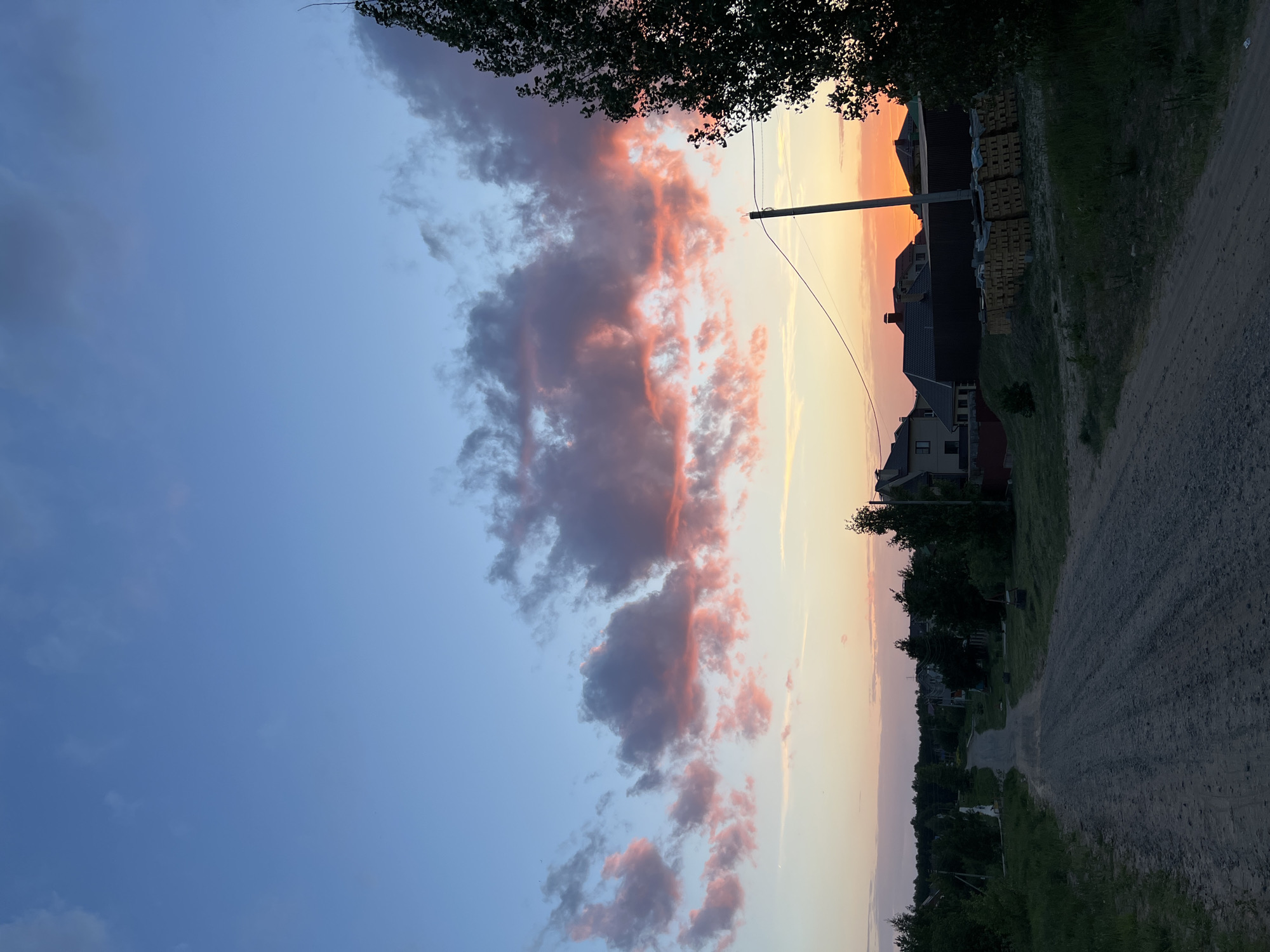 The image depicts a landscape at dusk or dawn in what appears to be a residential area. The sky is a gradient of blue to yellow and pink hues, with clusters of clouds tinged with pink. A utility pole stands vertically in the middle of the frame. The ground shows a dirt or gravel road lined with trees and vegetation. Residential buildings are visible along the roadside, with some piles of what may be bricks or construction materials near one of the houses. The image is vertically oriented. Based on the prompt and hints provided, it is located in the city of Noviy, Russia.
