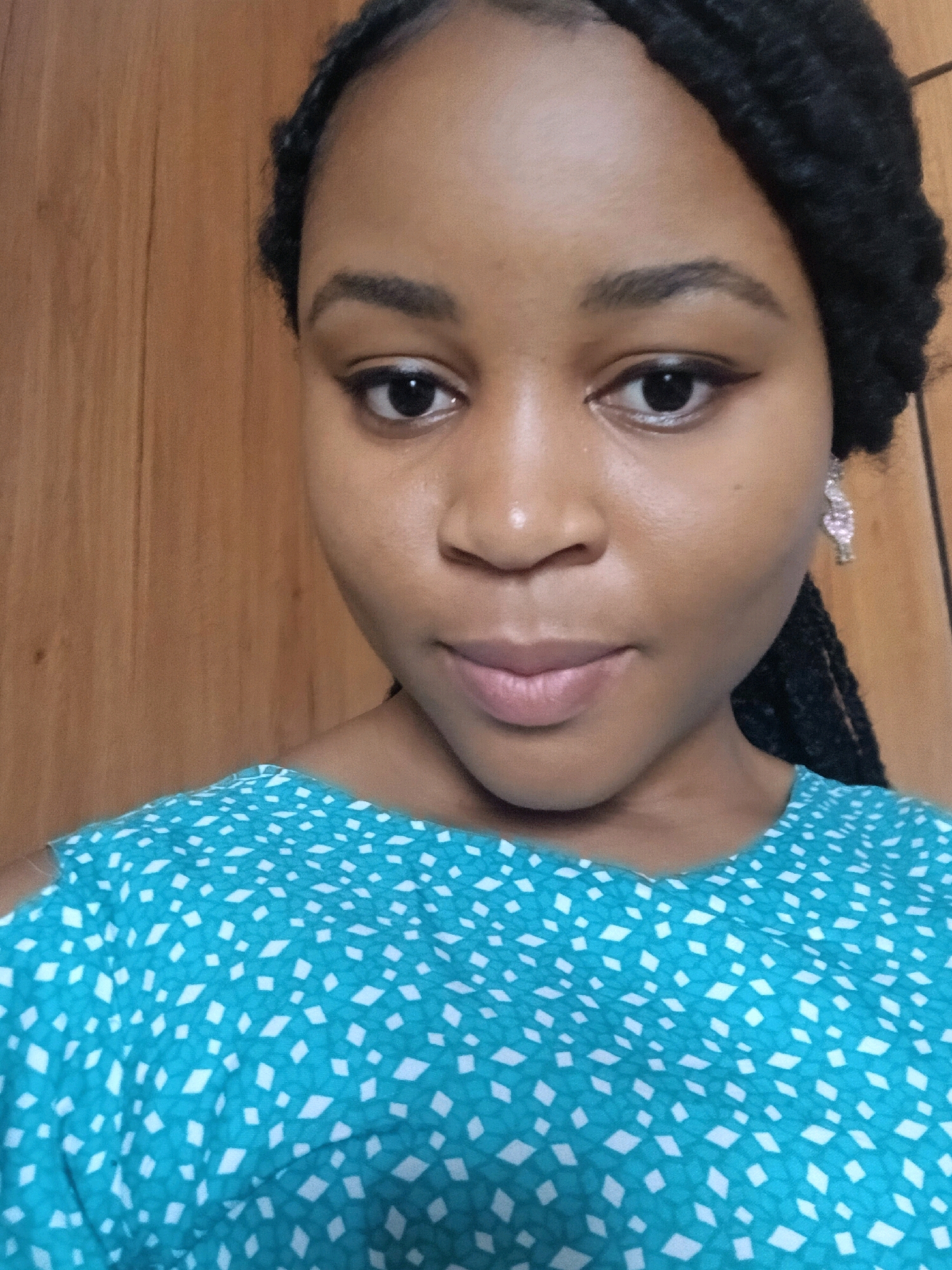 A young Black woman with braided hair and a turquoise and white patterned top is pictured against a wooden background. She is looking towards the camera. The image is a close-up shot, focusing on her face and upper body.