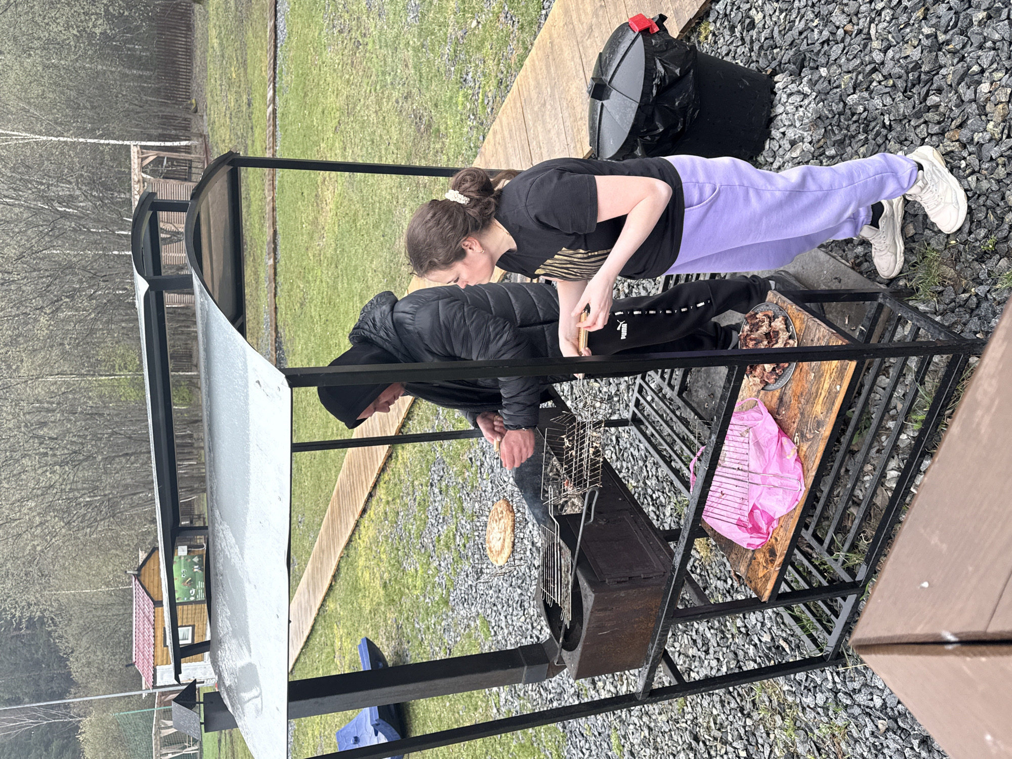 Two individuals are engaged in outdoor grilling at what appears to be a dacha or recreational area in Yekaterinburg, Russia. The scene captures a woman and a man preparing food on a metal BBQ structure with a roof, situated on a gray gravel ground interspersed with patches of grass.
The woman, with dark hair tied up, wears a black t-shirt with a gold/yellow design, light purple sweatpants, and white sneakers. She stands near a wooden surface on the grill structure, looking down intently, possibly attending to a plate of cooked meat, which resembles shashlik. The man, dressed in a black hooded puffer jacket, dark beanie, and black pants with a visible "Puma" logo, is actively grilling. He holds a grilling basket containing a round, flat food item (possibly bread or a patty) over the hot charcoal in the grill box.
The setting is an outdoor space featuring a wooden walkway or decking, sparse birch trees in the background, and distant wooden buildings. The overall ambiance suggests a cloudy or overcast day, as indicated by the soft lighting and the man's warm attire. A black trash can with a red lid and a pink plastic bag are also visible near the grilling area. The focus of the activity is clearly on preparing a meal outdoors, indicating a casual and relaxed gathering.