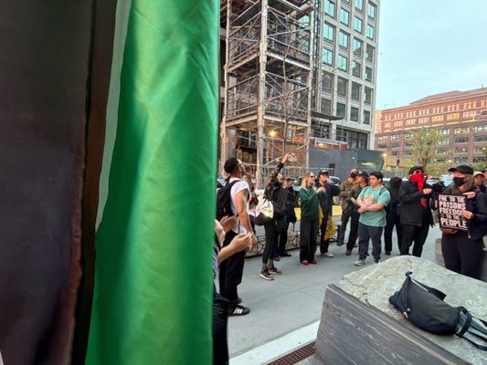 A photograph captures a group of individuals standing on a sidewalk in Hoboken, United States, in front of a building under construction. The group appears to be gathered for a demonstration or protest, with some individuals holding signs and flags. The environment includes urban elements such as buildings, scaffolding, and the sidewalk. The overall scene suggests an outdoor event with a focus on activism or public expression.