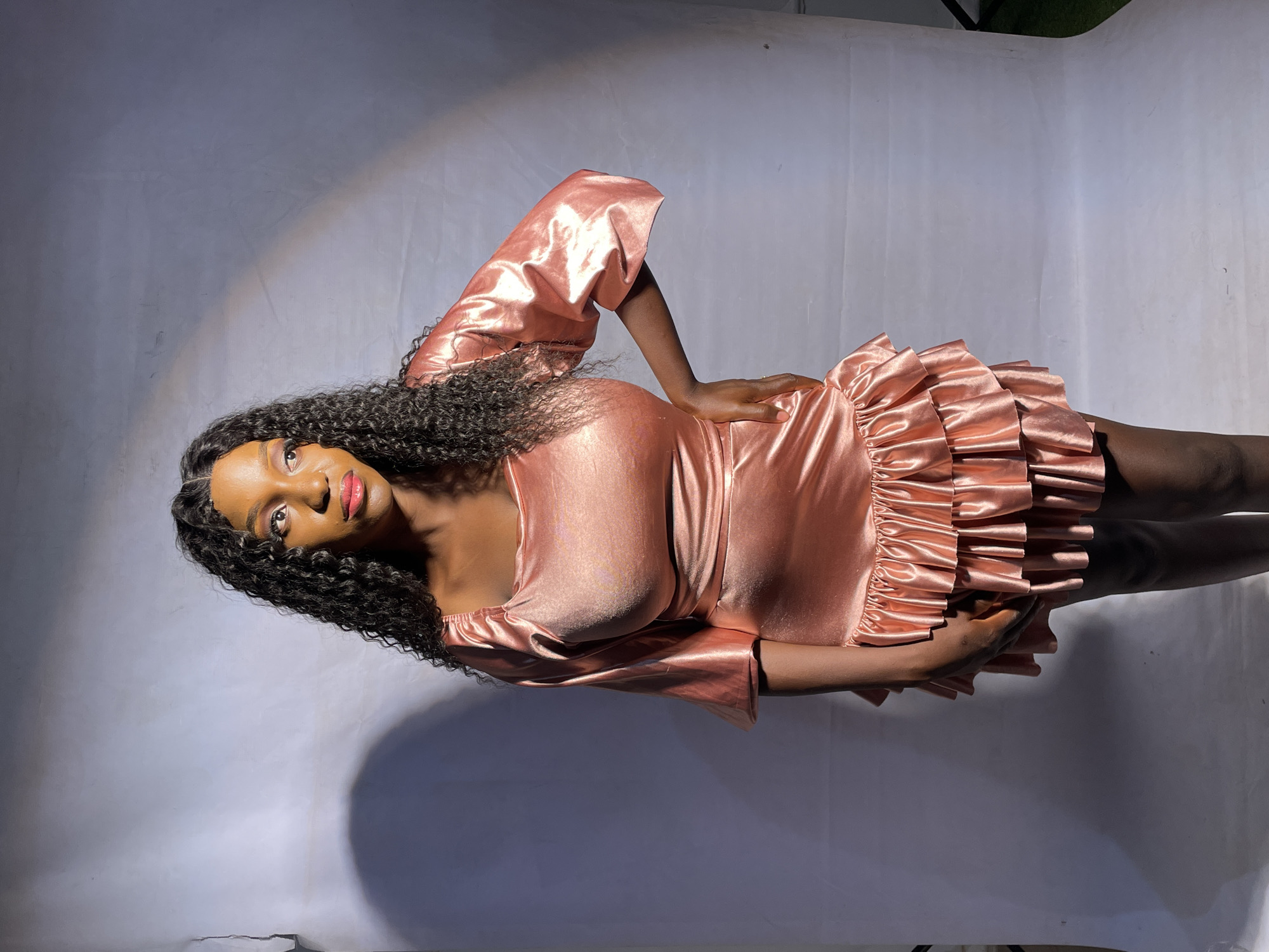 A young woman is lying on her side on a white surface. She has dark skin and long, curly black hair. She is wearing a light pink, short dress with ruffles around the hem. Her left arm is bent, with her hand resting on her waist. Her right arm is slightly bent, with her hand near her midsection. She is looking at the camera and smiling slightly. The lighting is uneven, with a bright spot near her head and a shadow cast across the background. The location is Barakin Akawo, Nigeria.