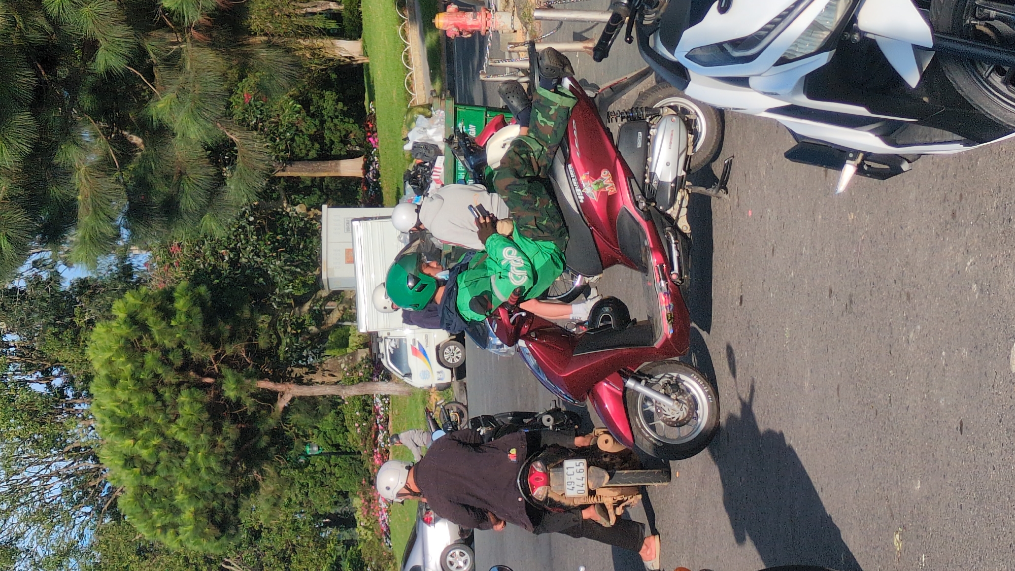 A street scene in Da Lat, Vietnam, features several individuals on motorcycles. In the foreground, a person wearing a green jacket with "Grab" branding and camouflage pants is seated on a maroon scooter, holding a mobile phone. To the left, another individual in a dark outfit and white helmet is on a different scooter. In the background, a white truck and several other people on motorcycles are visible. The environment includes trees, grassy areas, and a fire hydrant. The lighting suggests it is daytime.