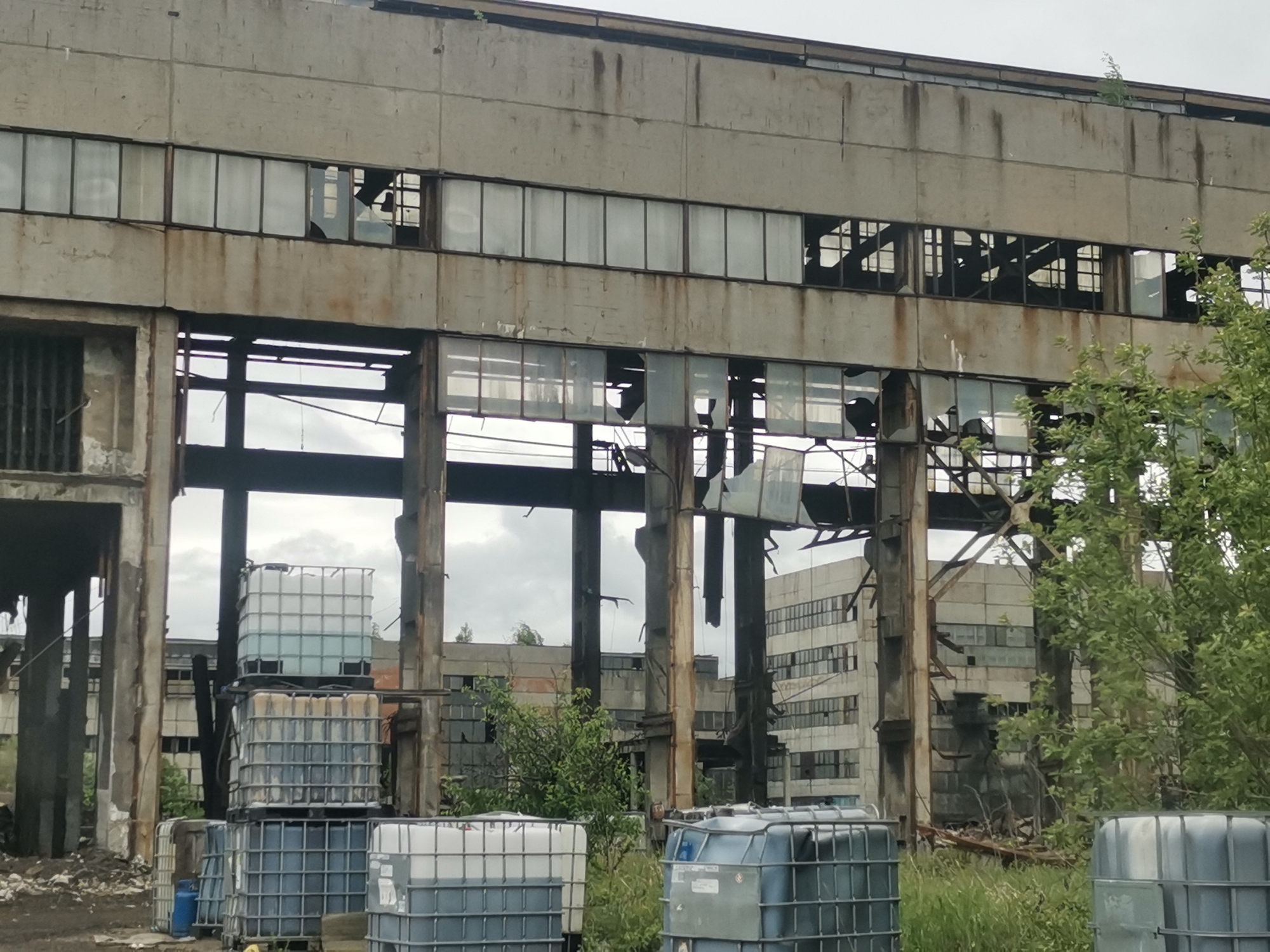 The image depicts an abandoned industrial building, likely located in an urban or semi-urban environment. The building exhibits significant decay, with broken windows and structural damage visible. The architecture appears to be utilitarian, typical of industrial structures. Several large plastic containers, possibly Intermediate Bulk Containers (IBCs), are present in the foreground, suggesting the site was once used for storage or processing of liquids. The surrounding environment shows signs of neglect, with overgrown vegetation and debris scattered around. In the background, another similar building can be seen. The sky is overcast. It cannot be confirmed that it is Zhivkovo, Bulgaria, based solely on the image.
