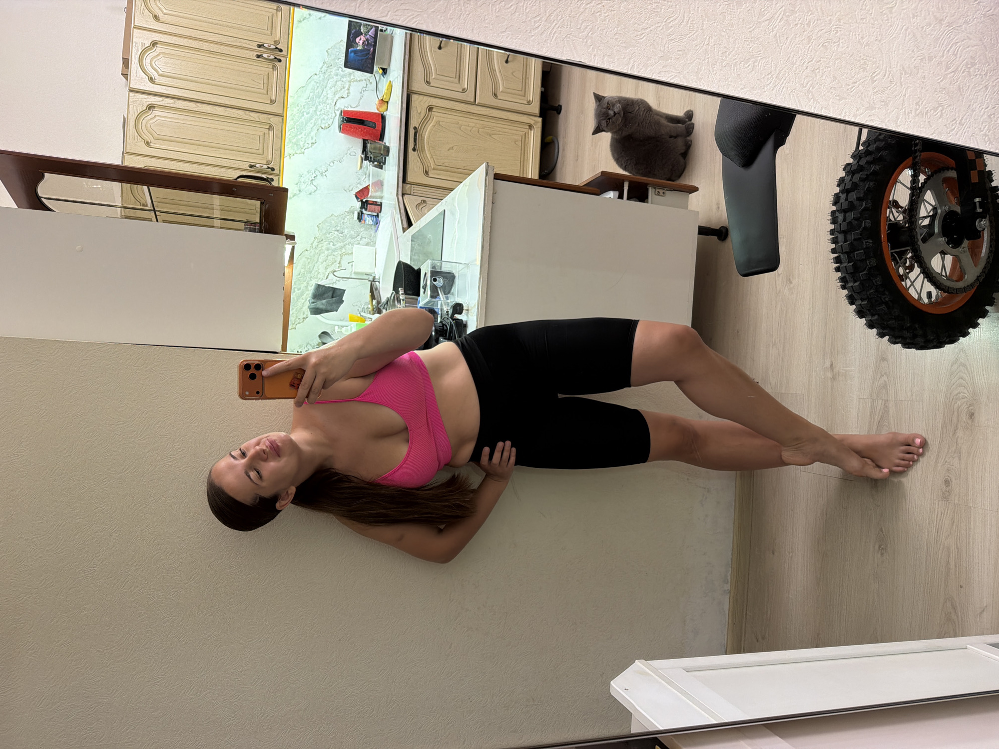 A woman takes a mirror selfie in what appears to be a residence in Sochi, Russia. The woman, with long dark hair, wears a pink ribbed sports bra and black cycling shorts, posing with her left hand on her hip and holding an orange-cased smartphone with her right hand. Her bare feet, showing pink painted toenails, are visible on a light wooden floor. The mirror's reflection reveals a kitchen with light-toned wooden upper and lower cabinets, a light countertop, a sink, and a patterned light green backsplash. Kitchen items visible on the counter include a red electric kettle, a laptop displaying a video of a man's face, and various containers. A gray cat sits on the wooden floor near the kitchen cabinets, looking towards the camera. In the lower right corner of the reflection, the rear wheel with an off-road tire and orange accents, along with part of the seat/fender of a small motorcycle or dirt bike, is visible. The wall behind the woman is a light beige with a subtle textured pattern.