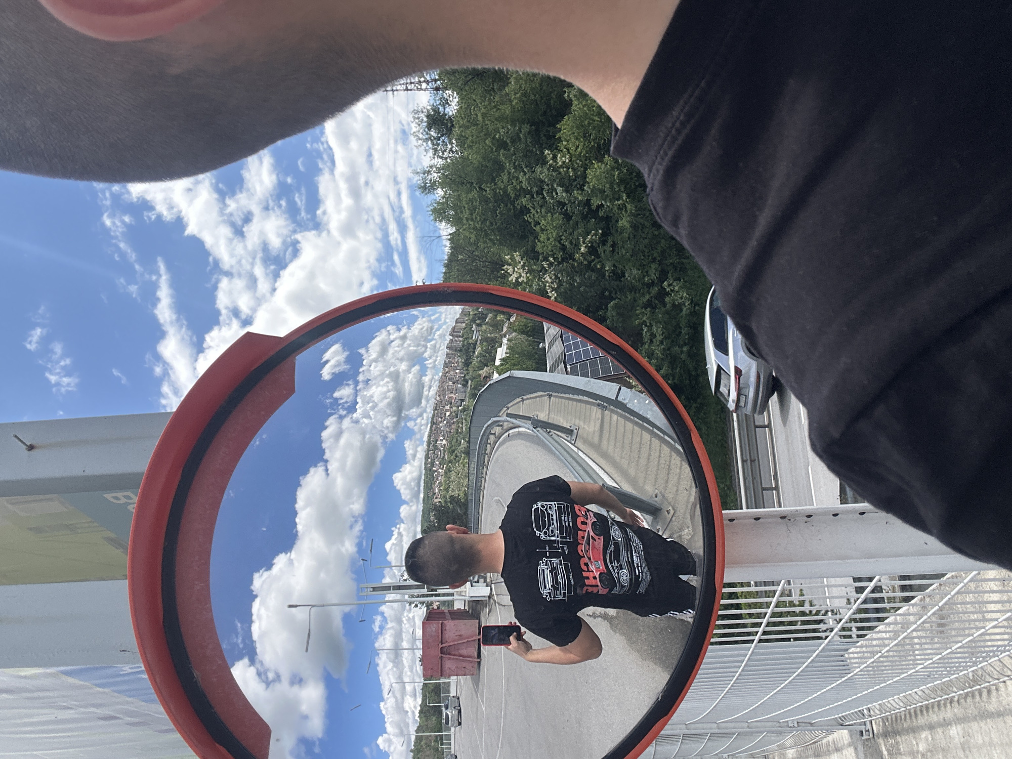 The image shows a fisheye reflection in a circular convex mirror. The mirror's orange frame is visible. A person wearing a black t-shirt with car graphics and black pants is seen holding a red smartphone within the reflection. The background in the reflection includes a blue sky with scattered clouds, a city skyline, and a gray pedestrian bridge. Parts of the photographer's head and shoulder are visible at the top and right of the frame. The mirror is mounted on a white metal railing. A car is visible out of focus. The scene is located on the pedestrian bridge over the train tracks in Veliko Tarnovo, Bulgaria, offering a view of the city and the surrounding landscape.