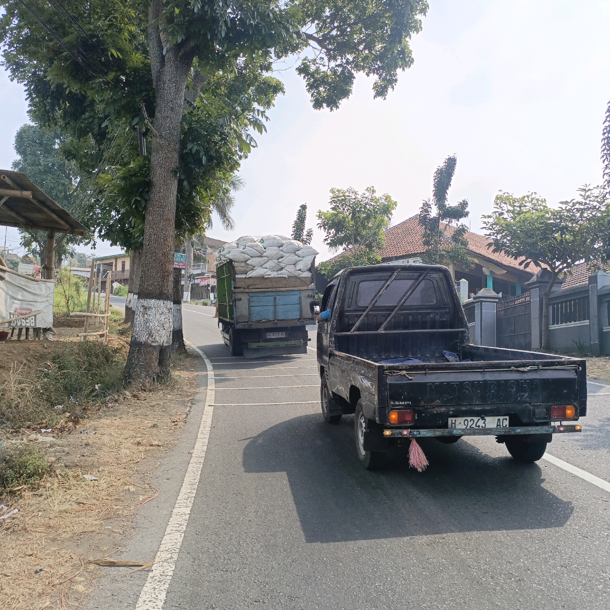 The image depicts a rural road scene in Sumogawe, Indonesia, under a bright, possibly overcast sky. In the foreground, a black Mitsubishi pickup truck, identified by its license plate "H-9243 AC", is traveling on the right side of the asphalt road. The truck has a metal cage structure in its bed and a pink tassel hanging from its rear bumper. The road is marked with a white line on the right edge. Ahead of the pickup, a green truck is visible, also traveling in the same direction, loaded with numerous white sacks. The truck has the license plate "AA-8662-VG".
To the left of the road, a large tree with thick bark, painted white in sections, stands prominently. Next to the tree, a small roadside shelter with a bamboo frame and a corrugated roof is visible. A white tarp covers part of the shelter. Dry grass and some litter are scattered on the ground along the roadside. In the background, houses with red tile roofs and fences are present. Green foliage from trees and plants surrounds the houses and lines the road. The overall scene suggests a typical day on a rural Indonesian road, with vehicles transporting goods.
